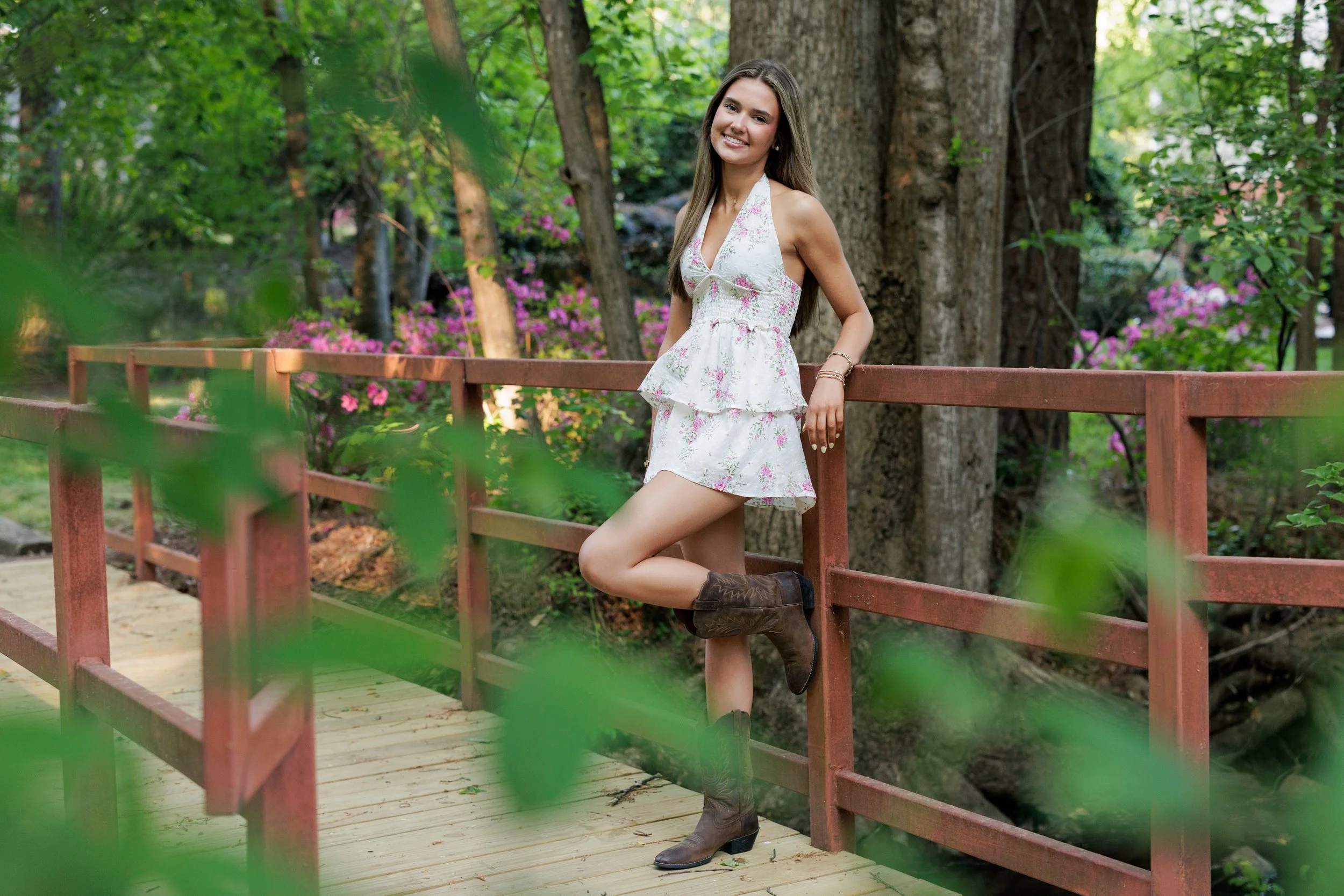 A young woman with long brown hair, wearing a white floral dress and cowboy boots, leaning against a wooden railing on a park bridge surrounded by green trees and pink flowers.