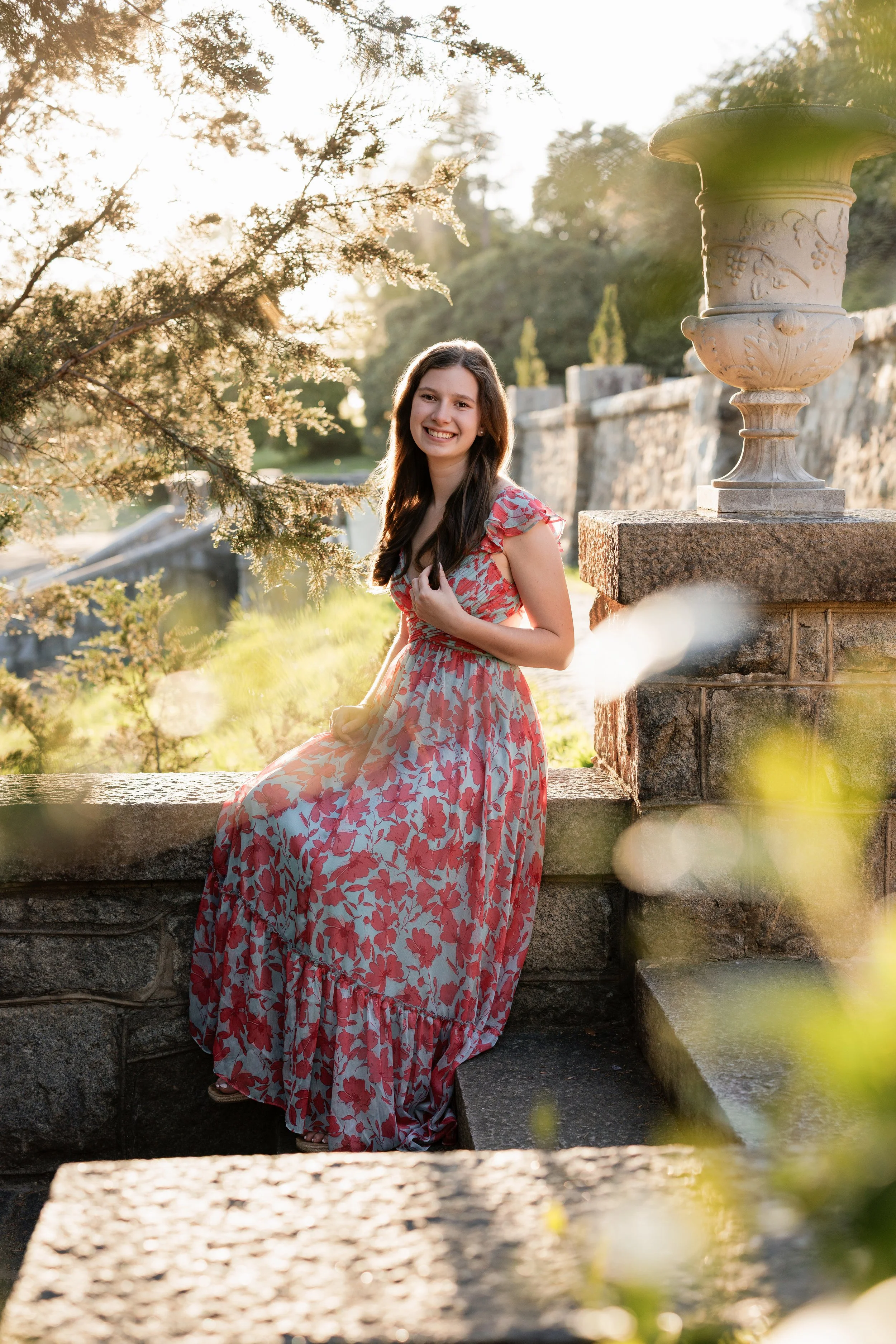 A young woman in a pink floral dress sitting on a stone ledge outdoors, smiling, with trees and a stone wall in the background during sunset.