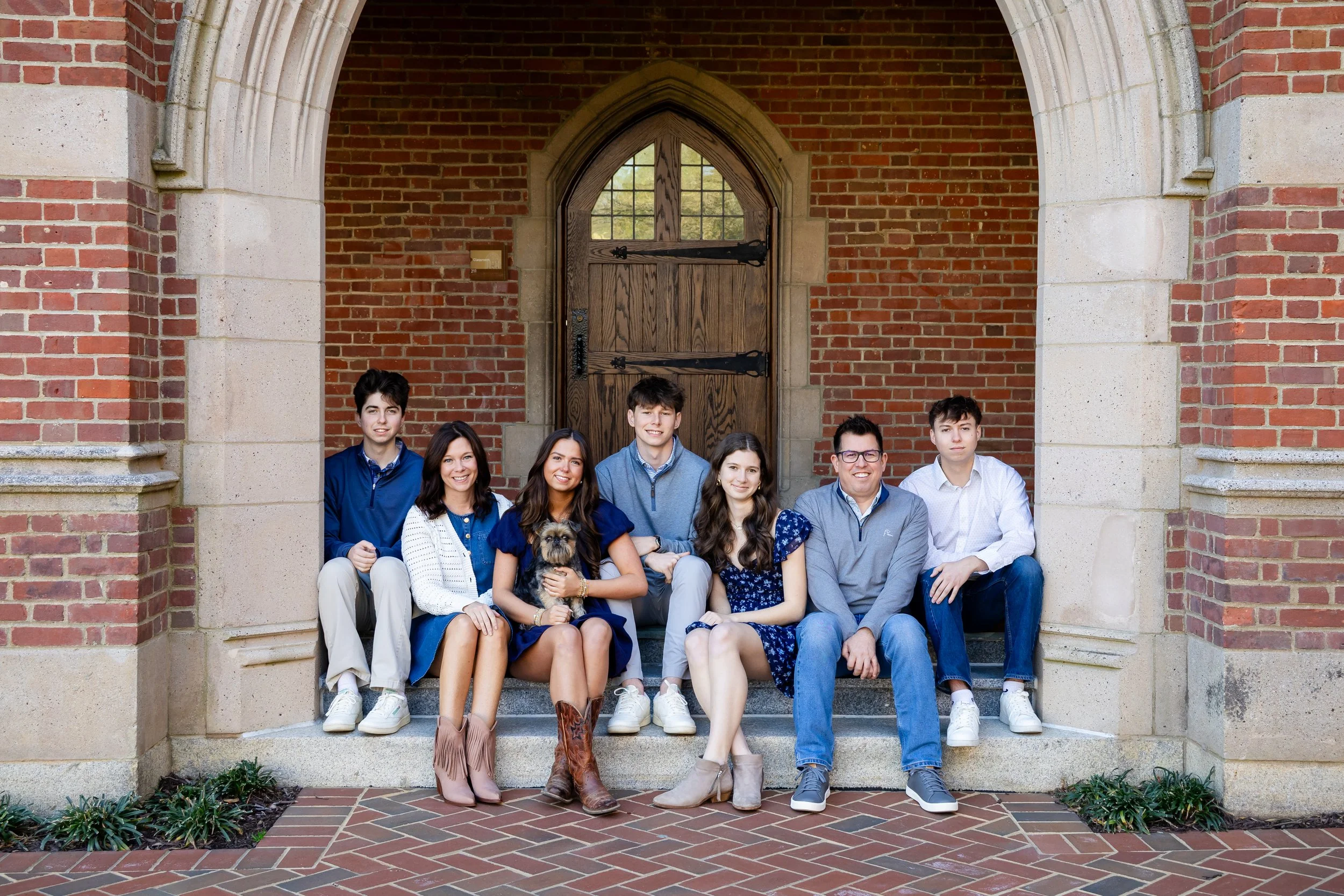A group of eight young people and a dog sitting on the steps in front of a brick building with a large wooden arched door.