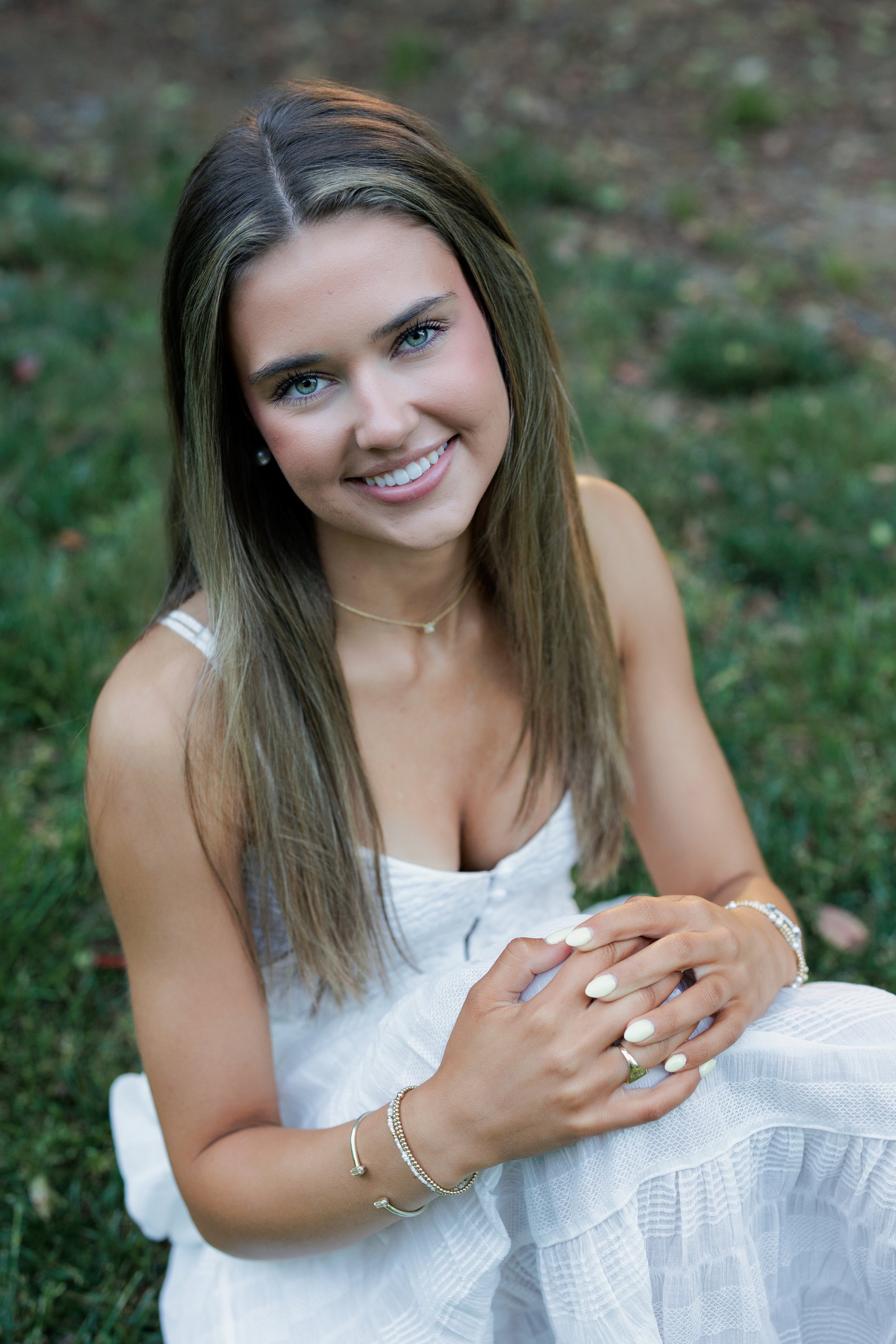 A young woman with long brown hair and blue eyes smiling, wearing a white dress, jewelry, and sitting outdoors on grass.