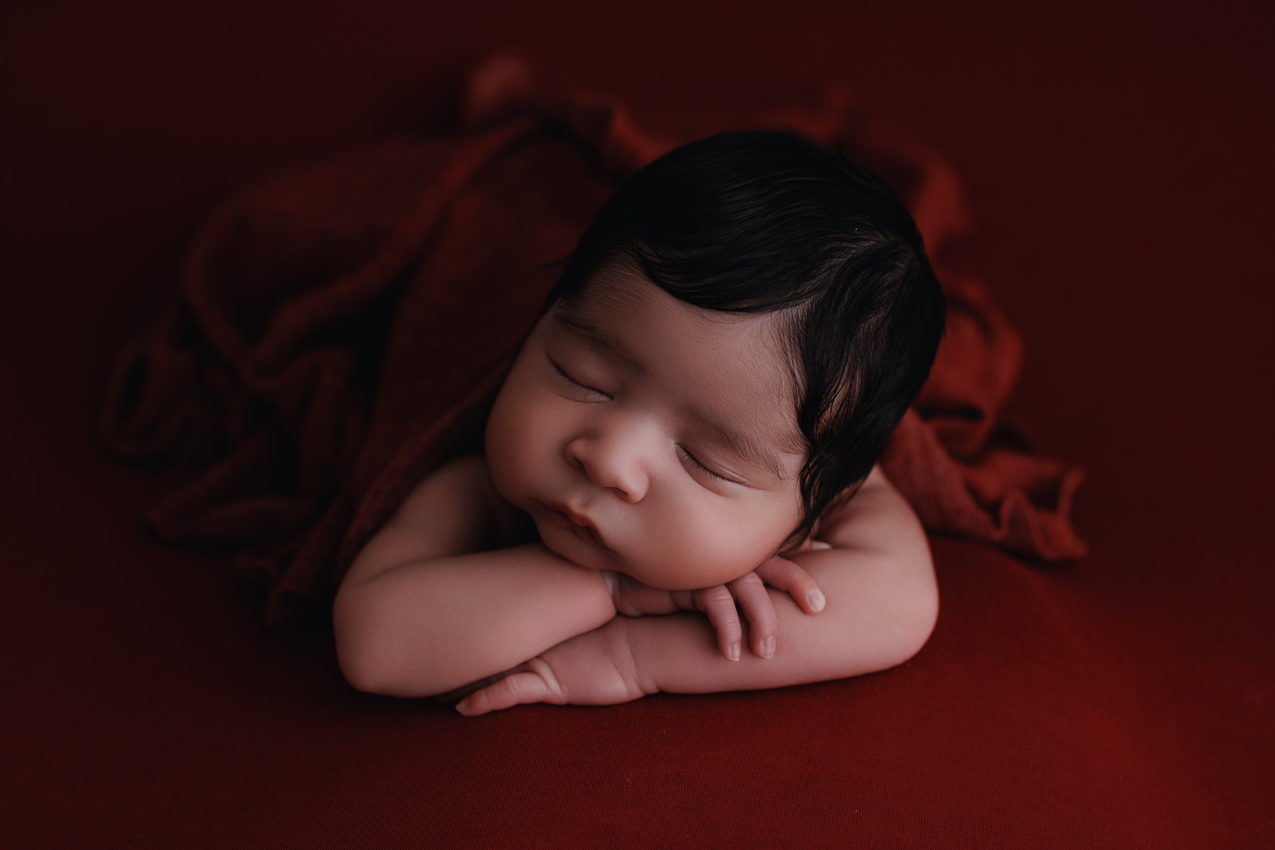 A sleeping infant with dark hair, resting on folded arms on a red background.