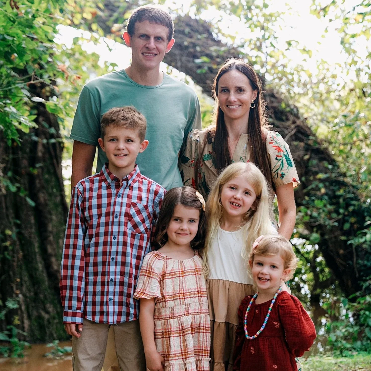 A family of eight standing outdoors in a wooded area, smiling at the camera. The group includes two adults and six children, all dressed in casual clothing.