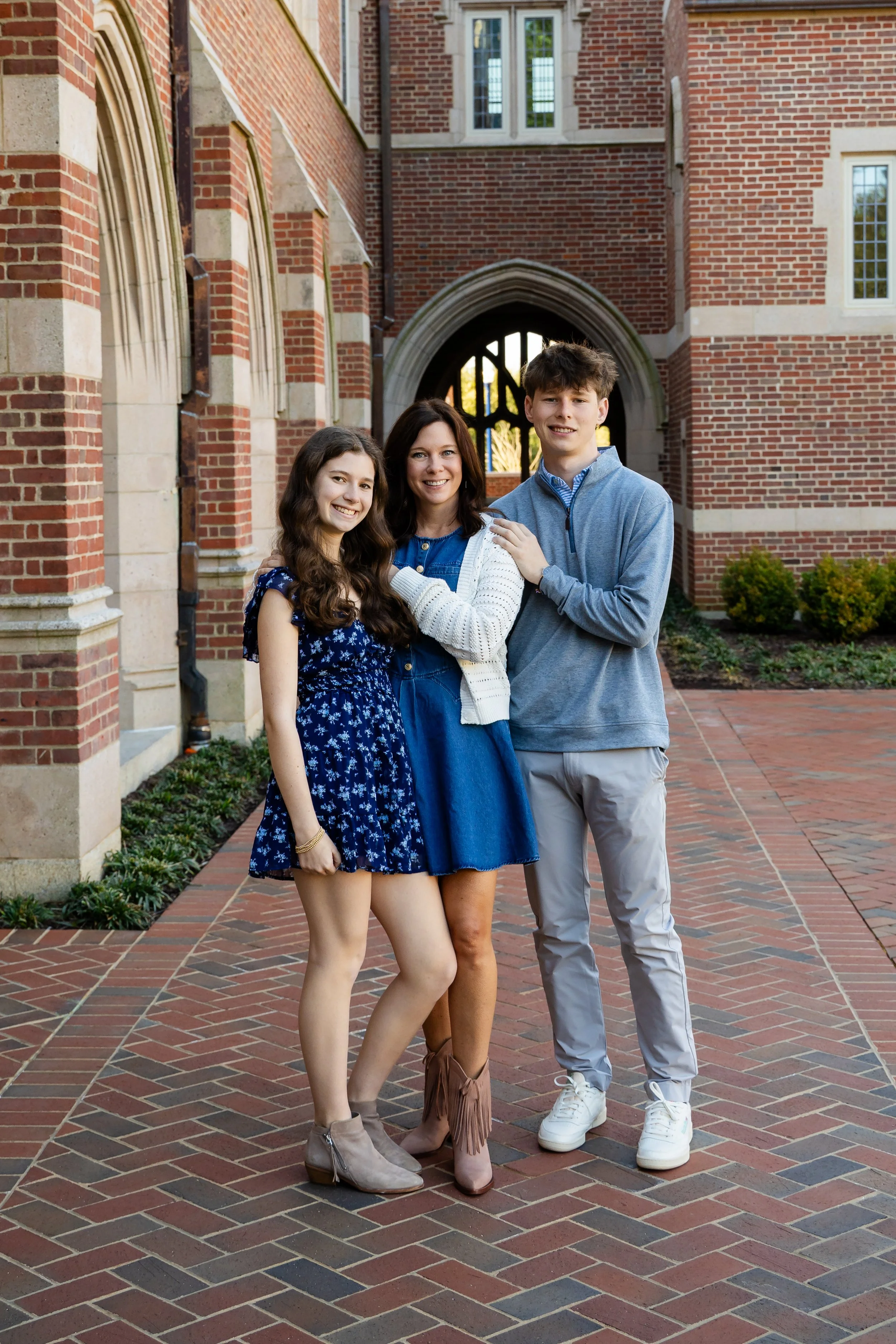 Three people, a young girl, a woman, and a young man, standing together in front of a red brick building with arched window and doorway, smiling at the camera.