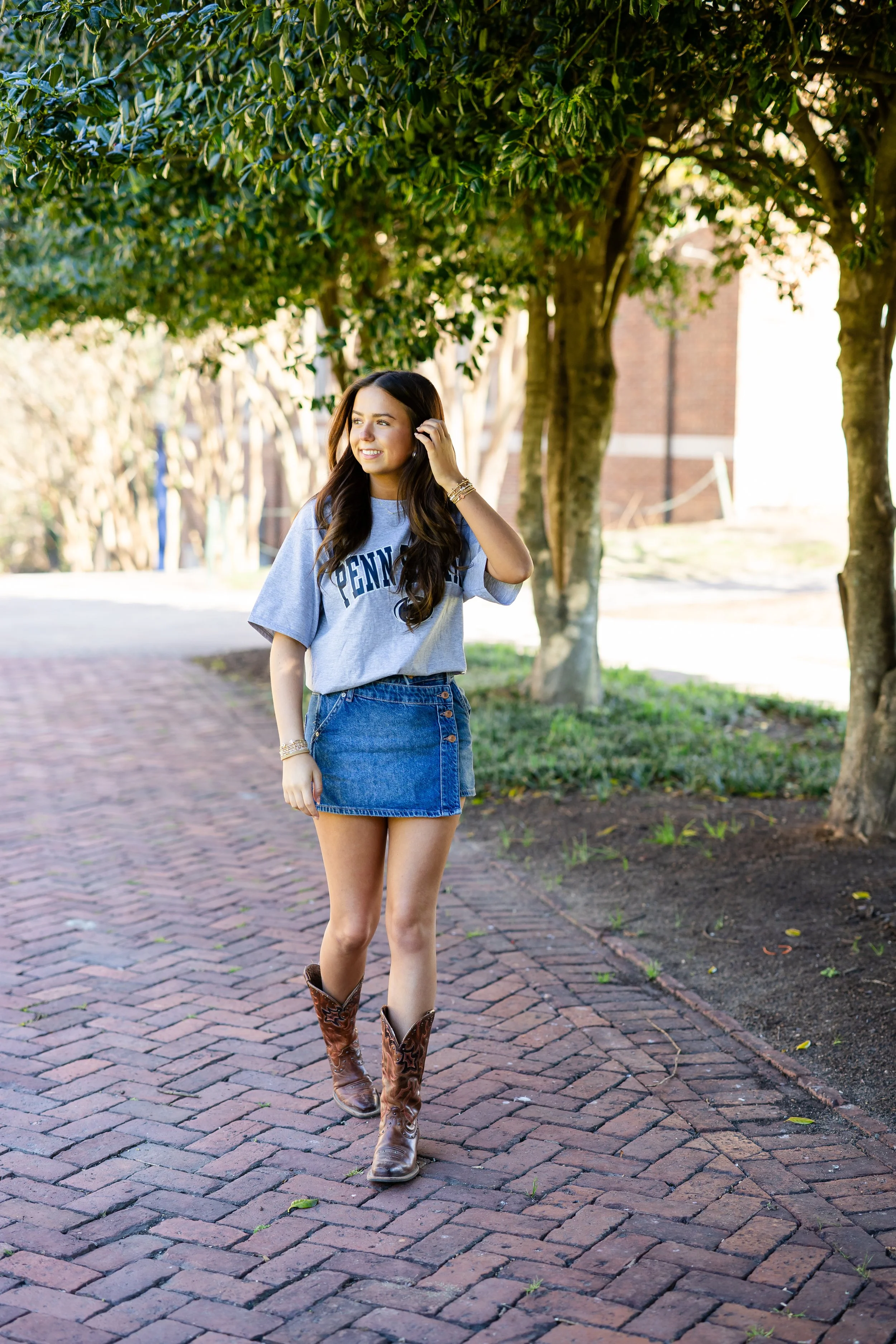 A young woman with long brown hair wearing a gray T-shirt, a denim skirt, and cowboy boots walking outdoors on a brick pathway, surrounded by trees and buildings in the background.