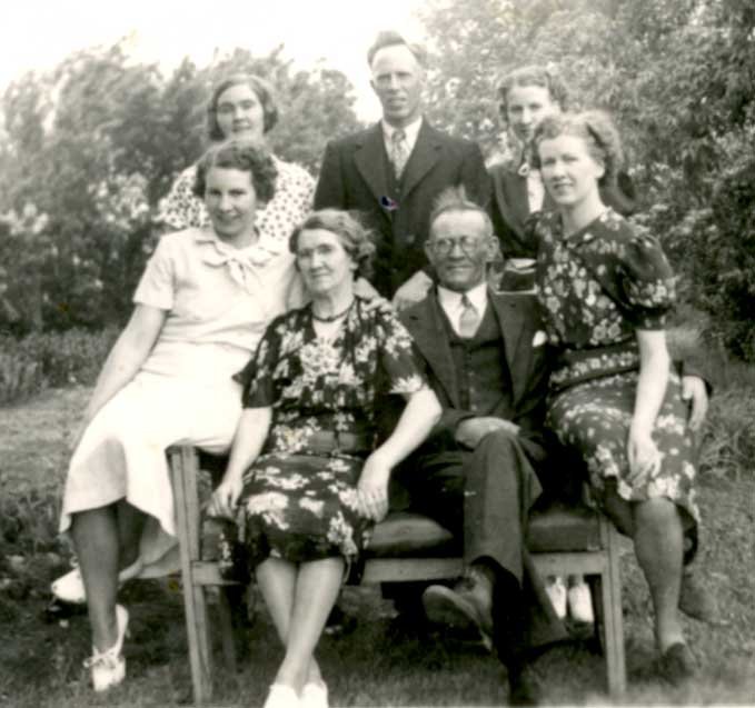  The Davidson family portrait in Manitoba. Older parents sit on garden bench surrounded by children. Four daughters and one son.  