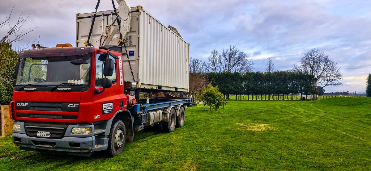 A red flatbed truck carrying a white shipping container parked on a grassy field with trees and a cloudy sky in the background.