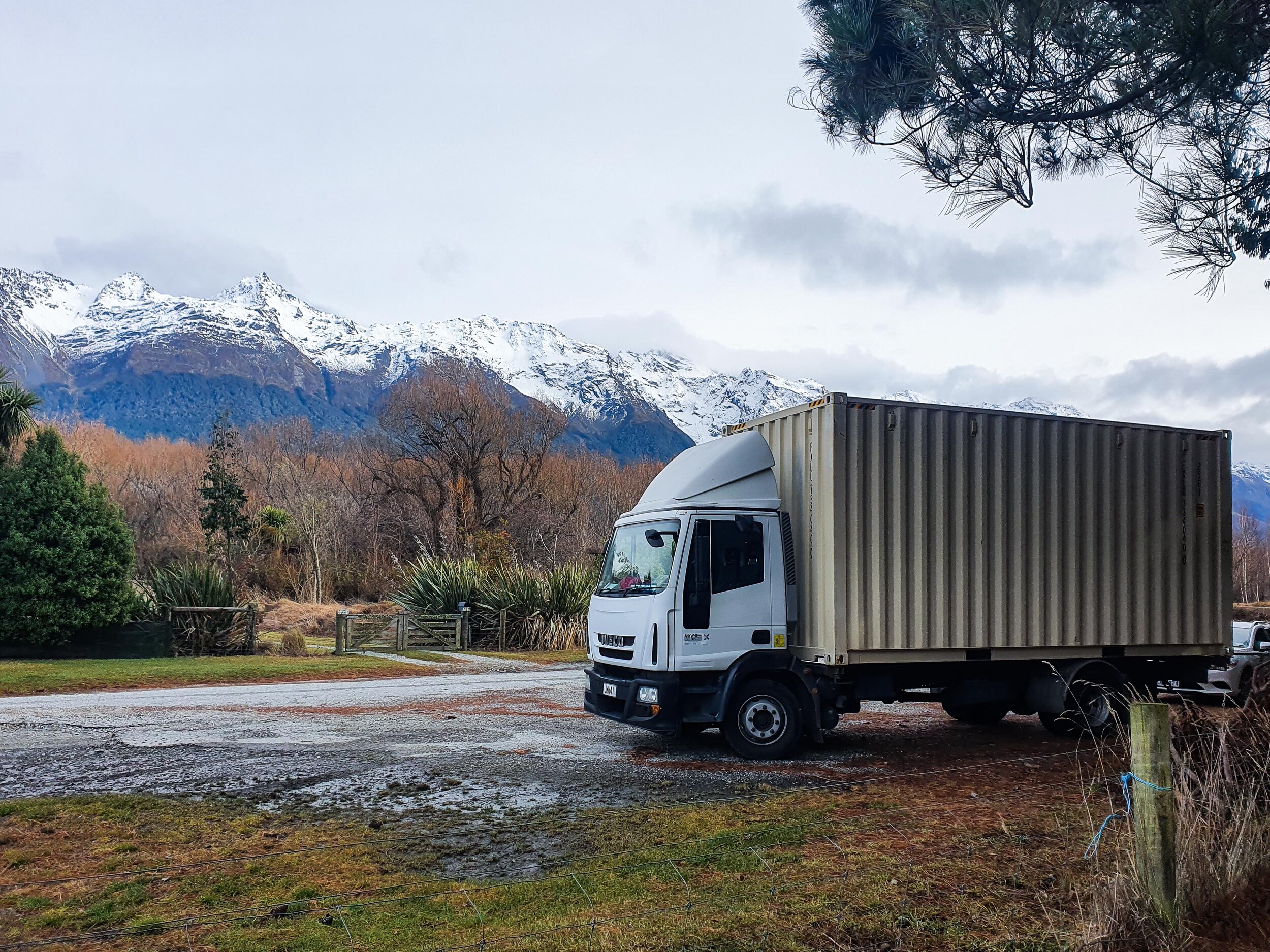 Truck parked on a gravel path with snowy mountains in the background and trees around.
