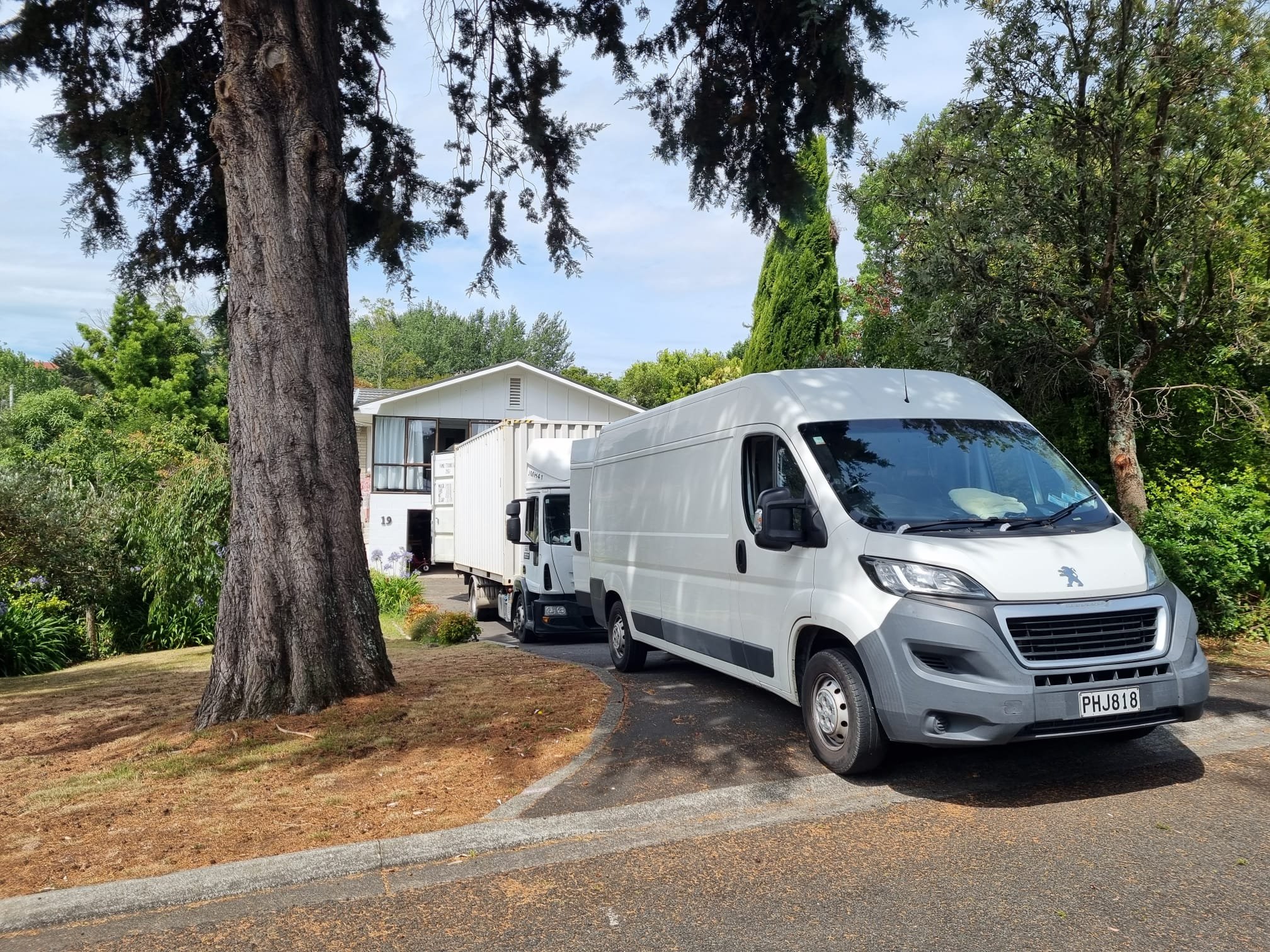 Moving trucks parked outside a house surrounded by trees and greenery.