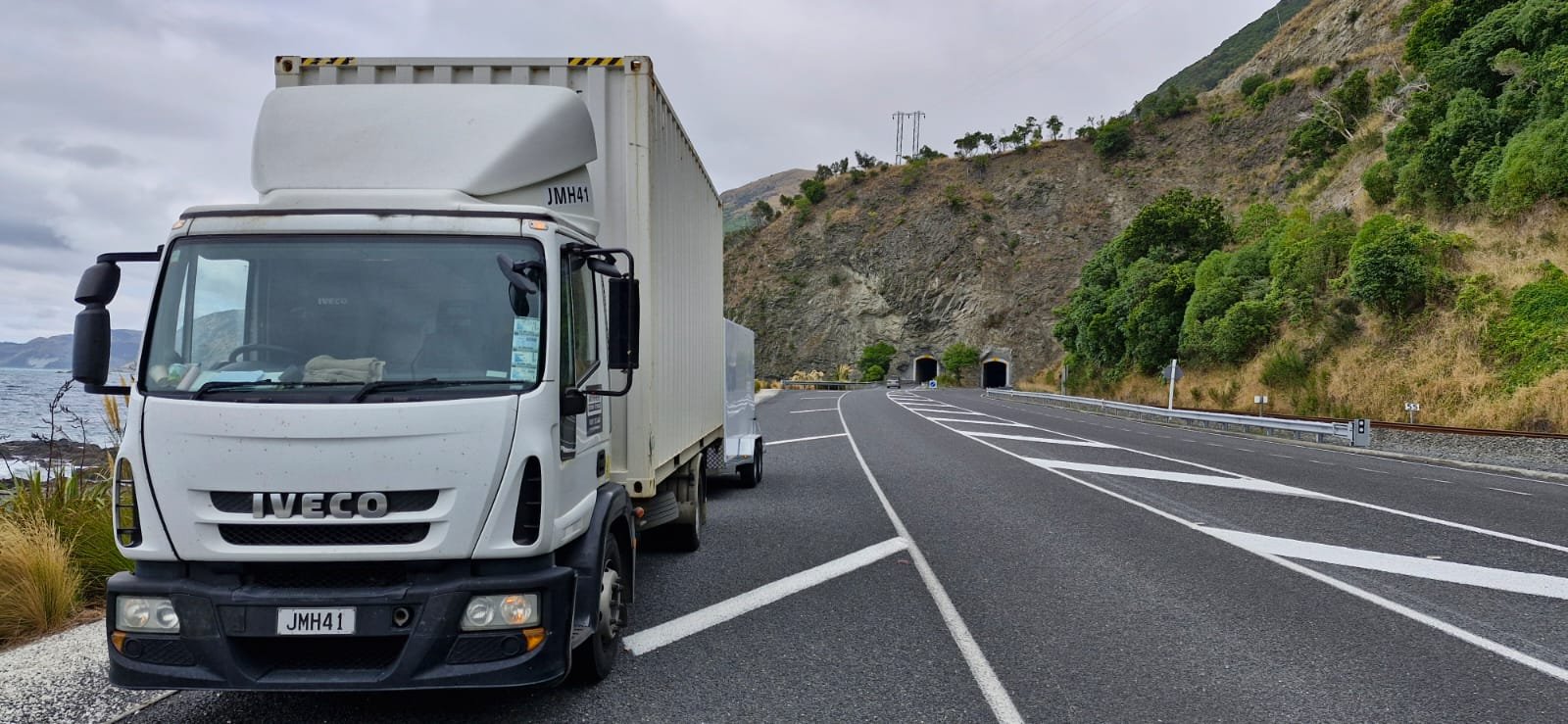 A white truck parked on a highway near a mountainous area with tunnels, on a cloudy day.