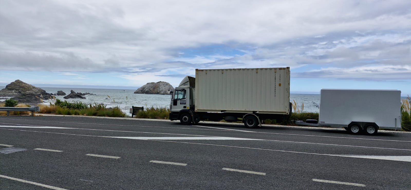 Truck and trailer parked by the ocean with rocky shoreline and cloudy sky in the background.