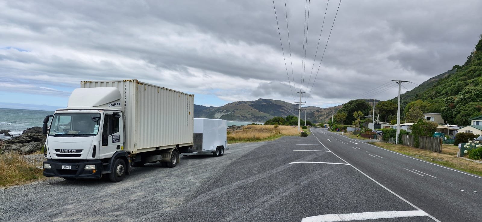 A white truck with a container parked on a roadside near the ocean, mountains in the background, overcast sky.