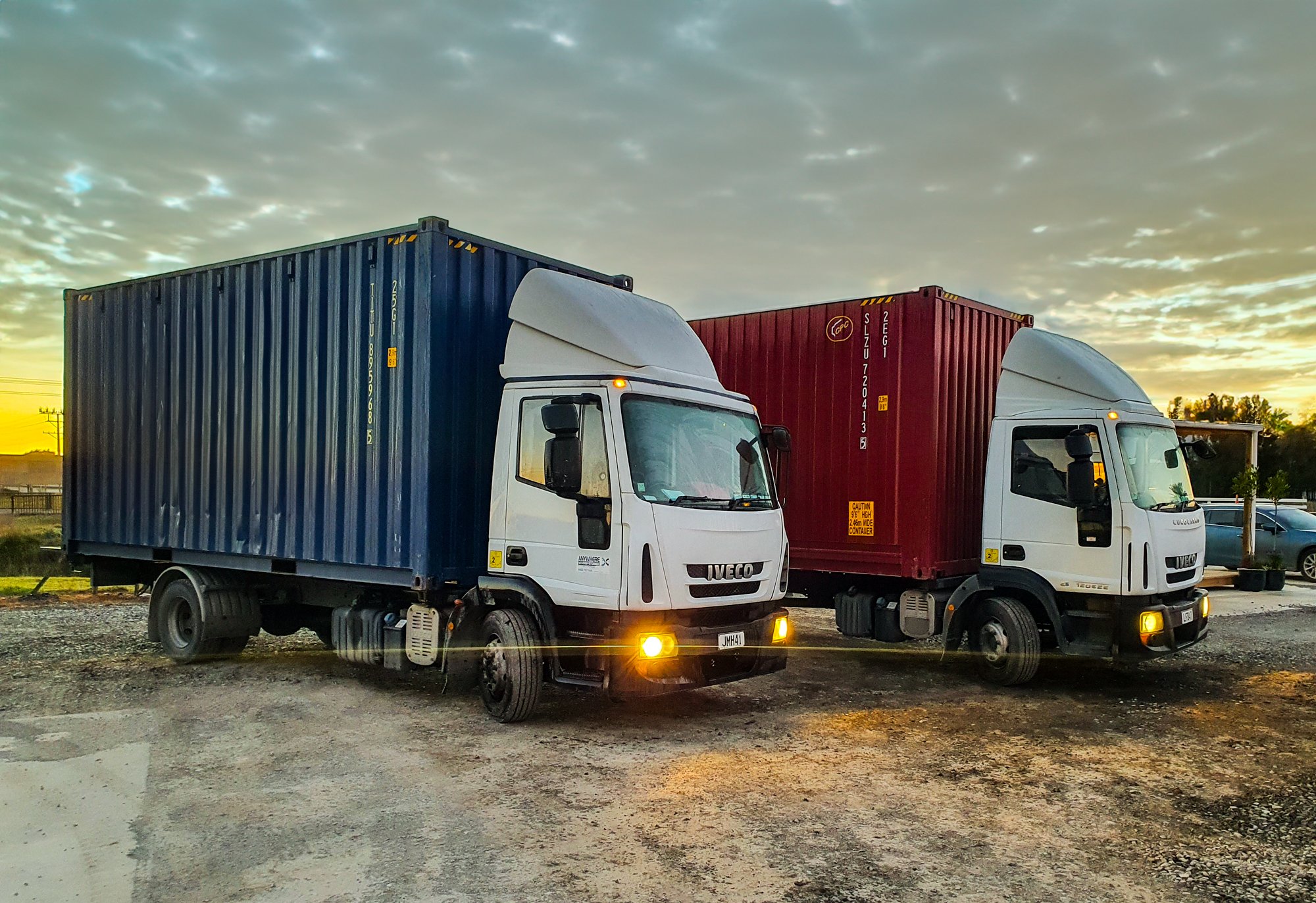 Two white Iveco trucks with shipping containers, one blue and one red, parked on gravel at sunset.