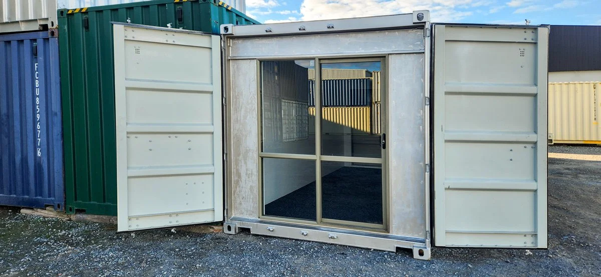A shipping container with open sliding glass doors, next to others in a storage yard.