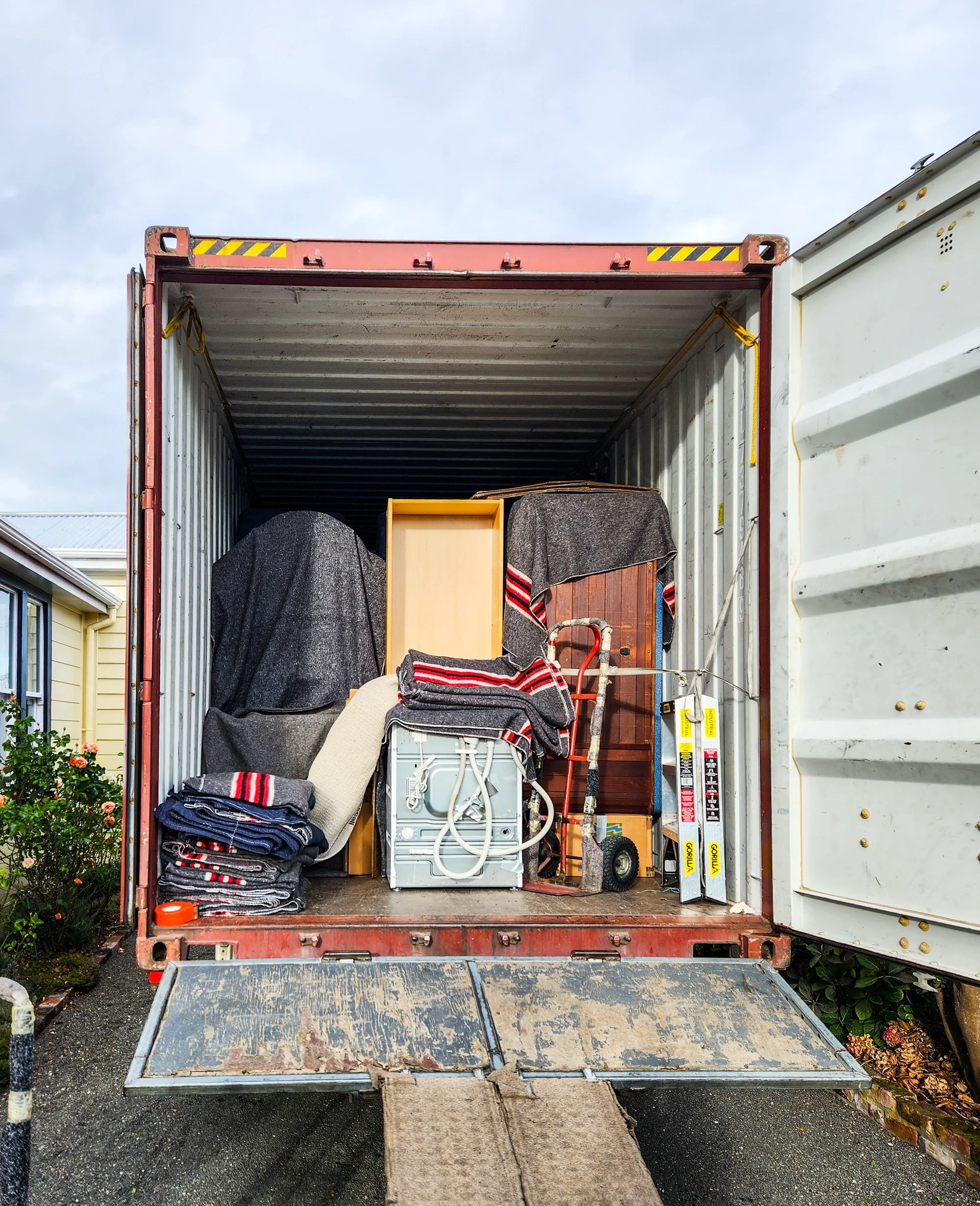 Opened moving truck filled with wrapped furniture, boxes, and household items, parked outside near a yellow house and greenery.