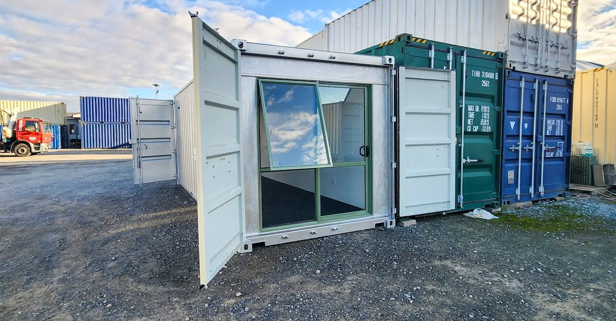 A white shipping container with glass doors and windows, situated among other cargo containers in an outdoor lot.