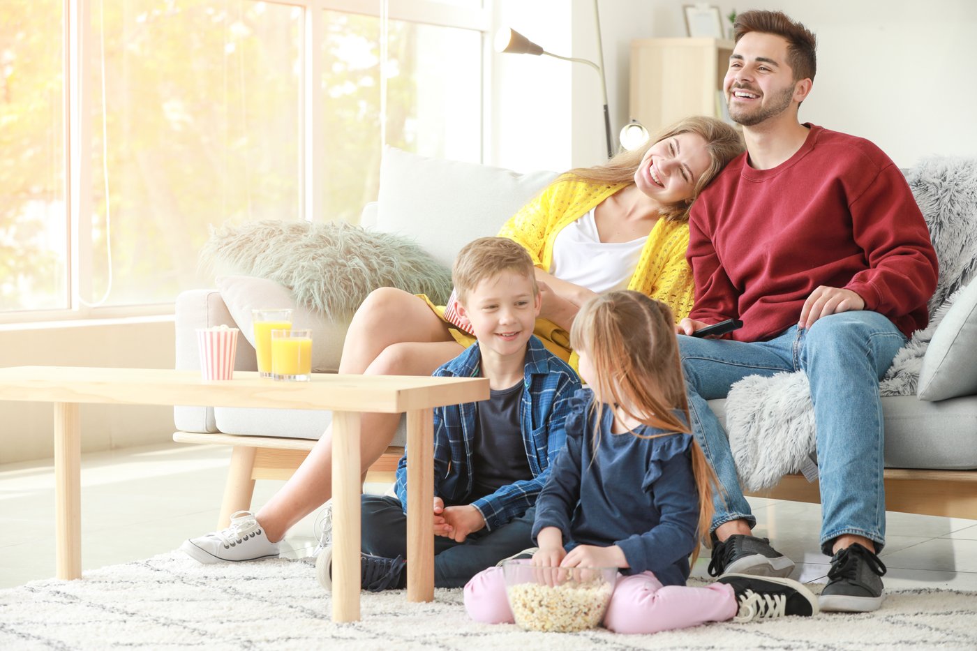 A family of four enjoying time together in a living room. The parents are sitting on a couch while two children sit on the floor in front of a coffee table with popcorn and drinks. The room is bright and cozy.