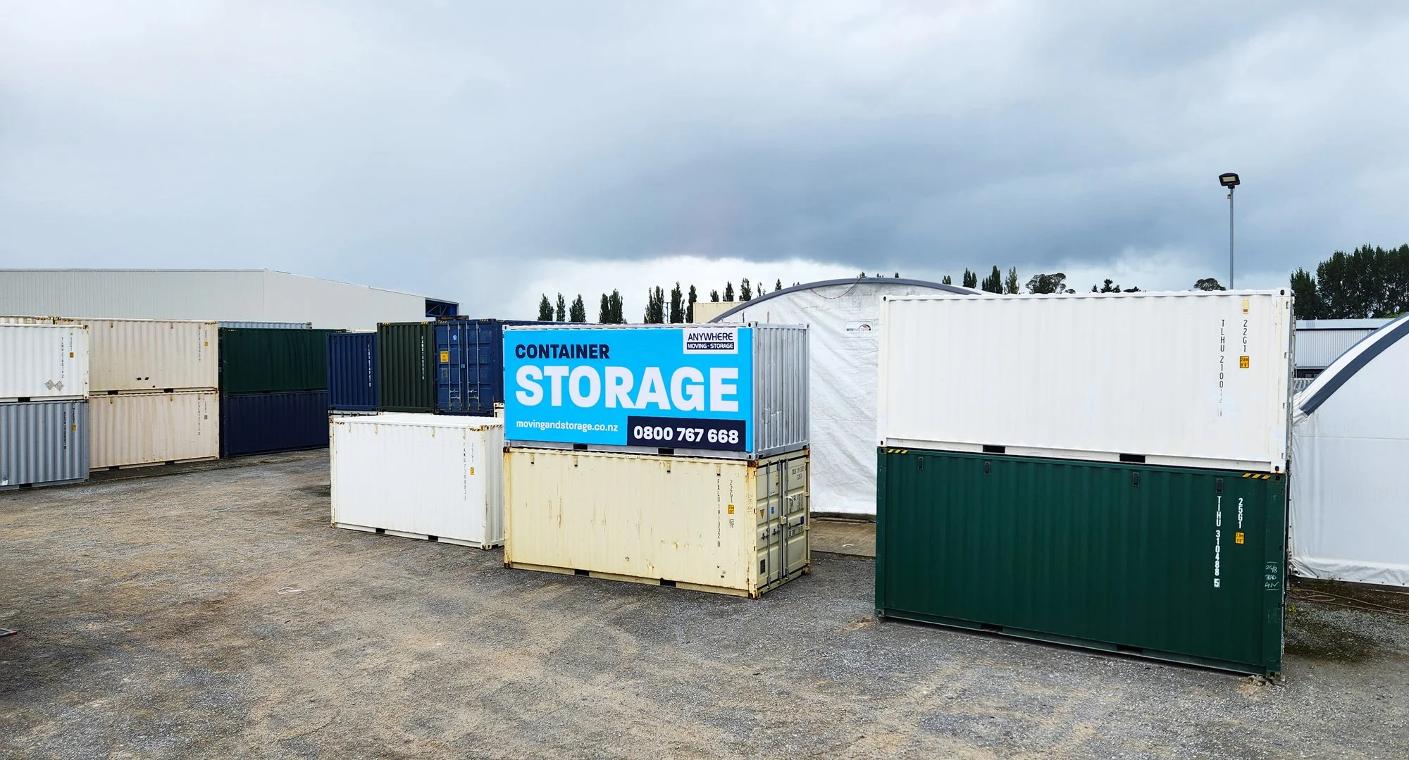 Outdoor storage lot with multiple shipping containers in different colors, including white, green, beige, and blue, some with logos and identification numbers, under an overcast sky.