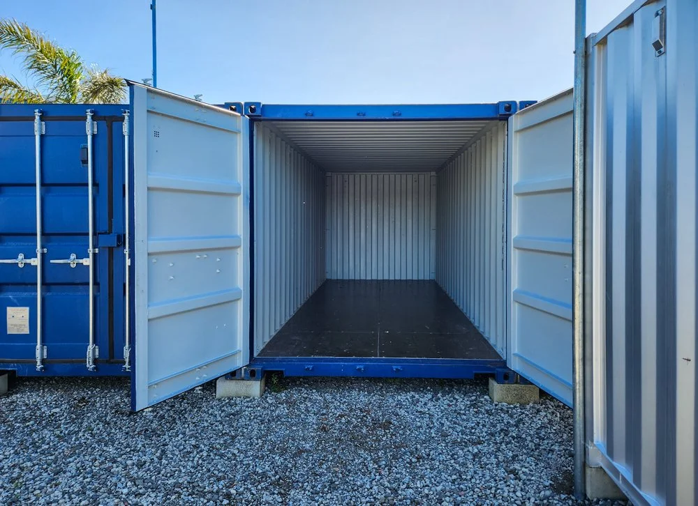 Empty blue shipping container with open doors, situated on gravel ground outdoors against a background of palm trees and blue sky.
