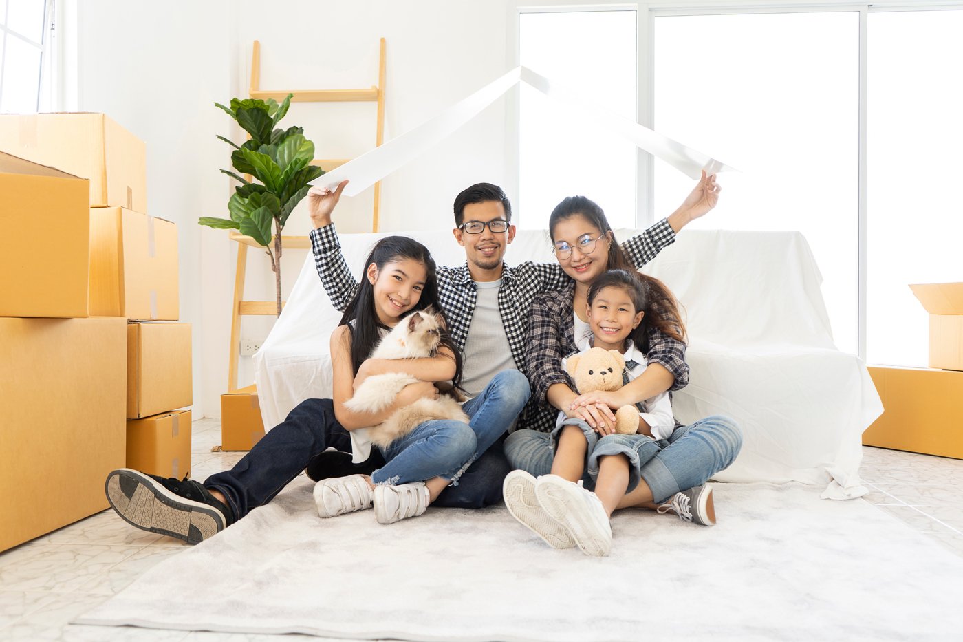 Family sitting together on the floor in a new home surrounded by moving boxes. The father holds a paper roof above them, symbolizing a house, while the daughter holds a cat and the other child a teddy bear. A potted plant and ladder shelf are in the background.
