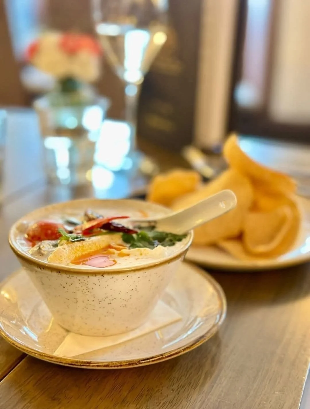 A bowl of Asian soup with vegetables and a fried egg, served on a beige plate with sprigs of basil, placed on a wooden table. In the background, there is a plate with crispy potato chips.