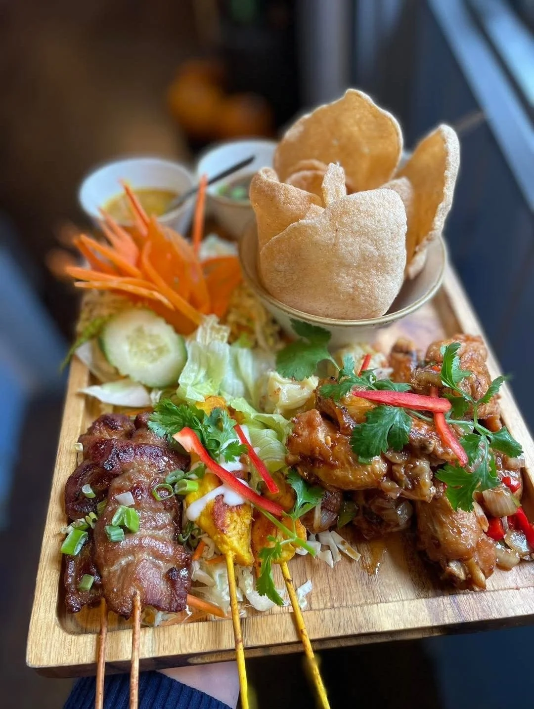 A wooden platter with grilled skewers, fresh vegetables, fried wontons, and garnished with cilantro and red pepper slices.