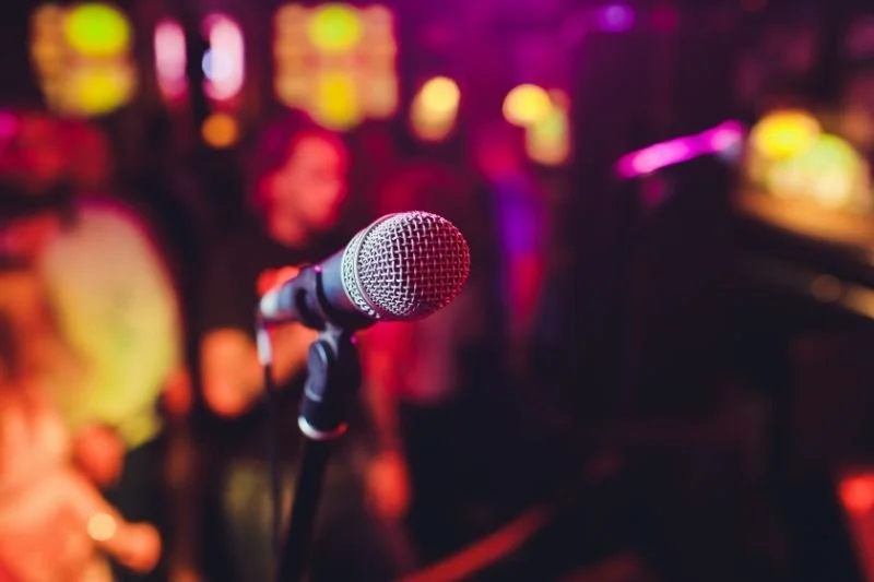 Close-up of a microphone on a stand with a crowd in the background at a lively indoor music venue or nightclub.