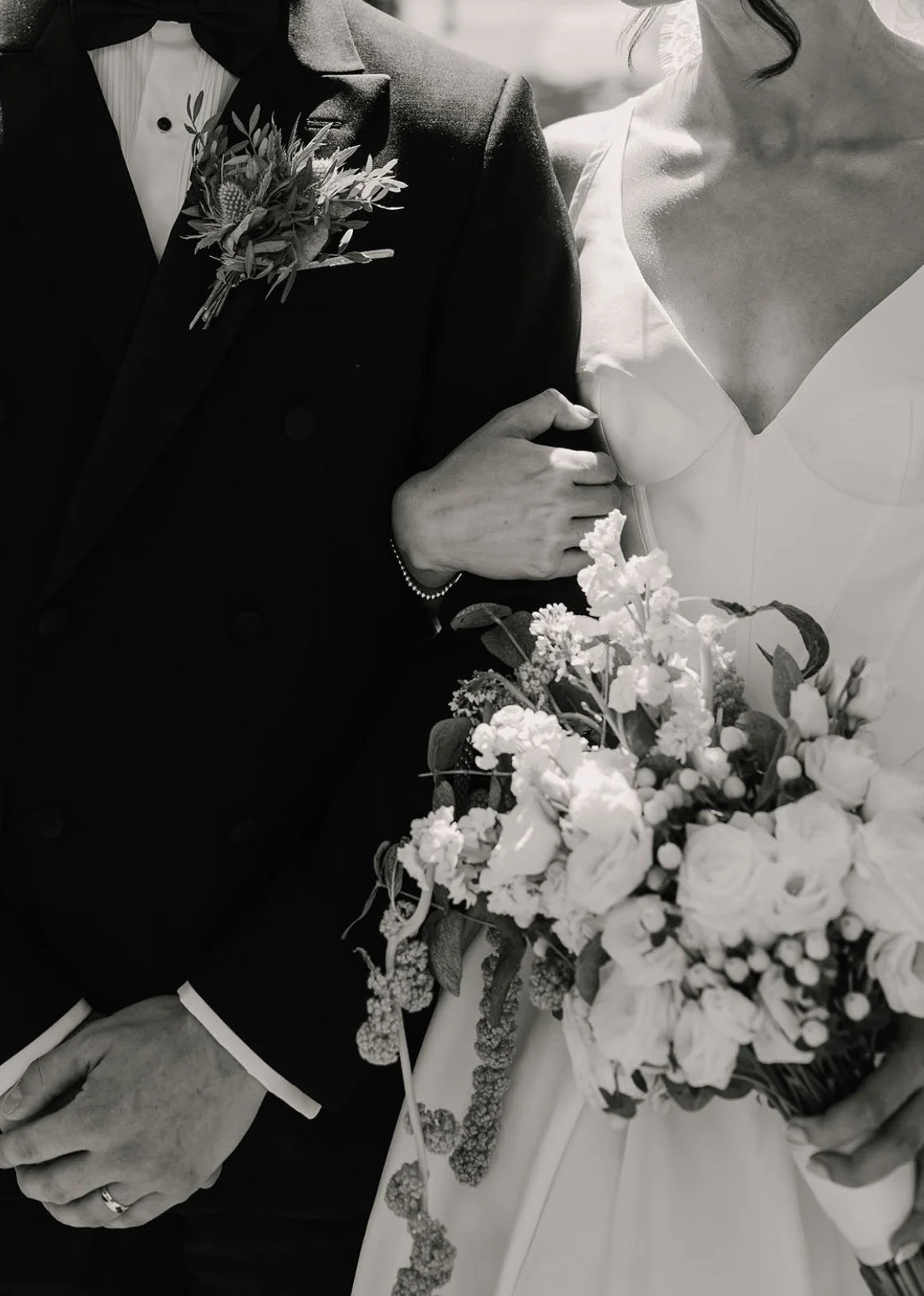 Close-up of a couple in wedding attire, with the bride holding a bouquet of flowers and the groom wearing a boutonniere, both holding hands.