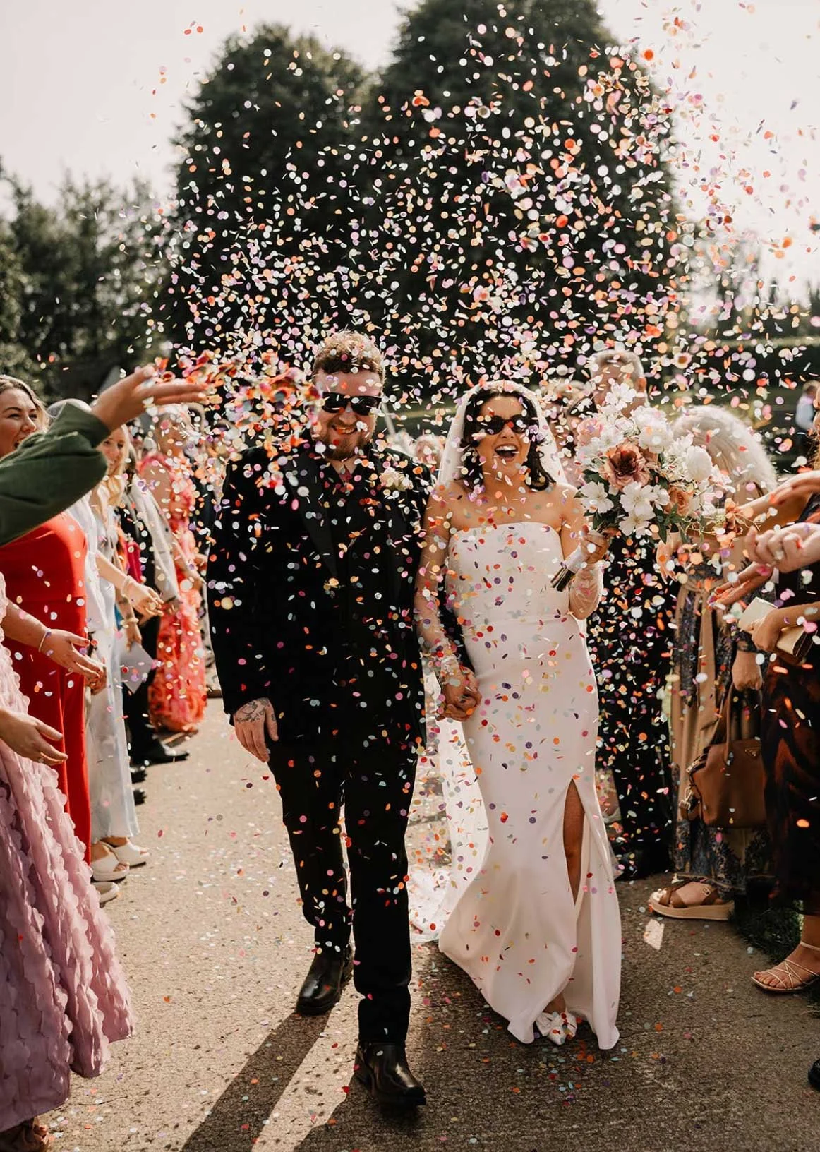 A bride and groom holding hands and walking through a crowd of people as confetti falls around them at their wedding celebration outdoors.
