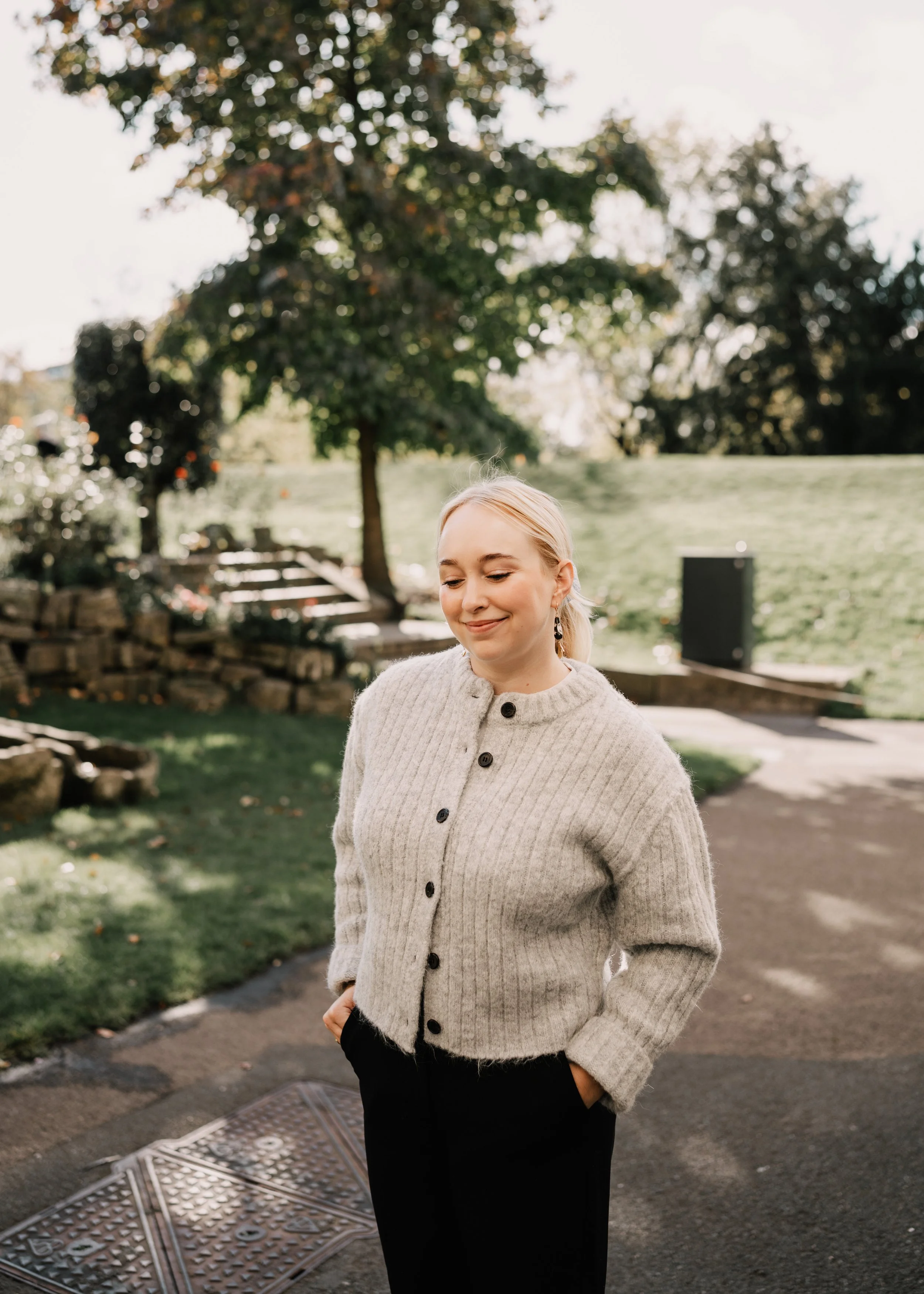 A woman with blonde hair, dressed in a beige cardigan with black buttons, standing outdoors on a sidewalk with greenery and trees in the background, smiling with her eyes closed.