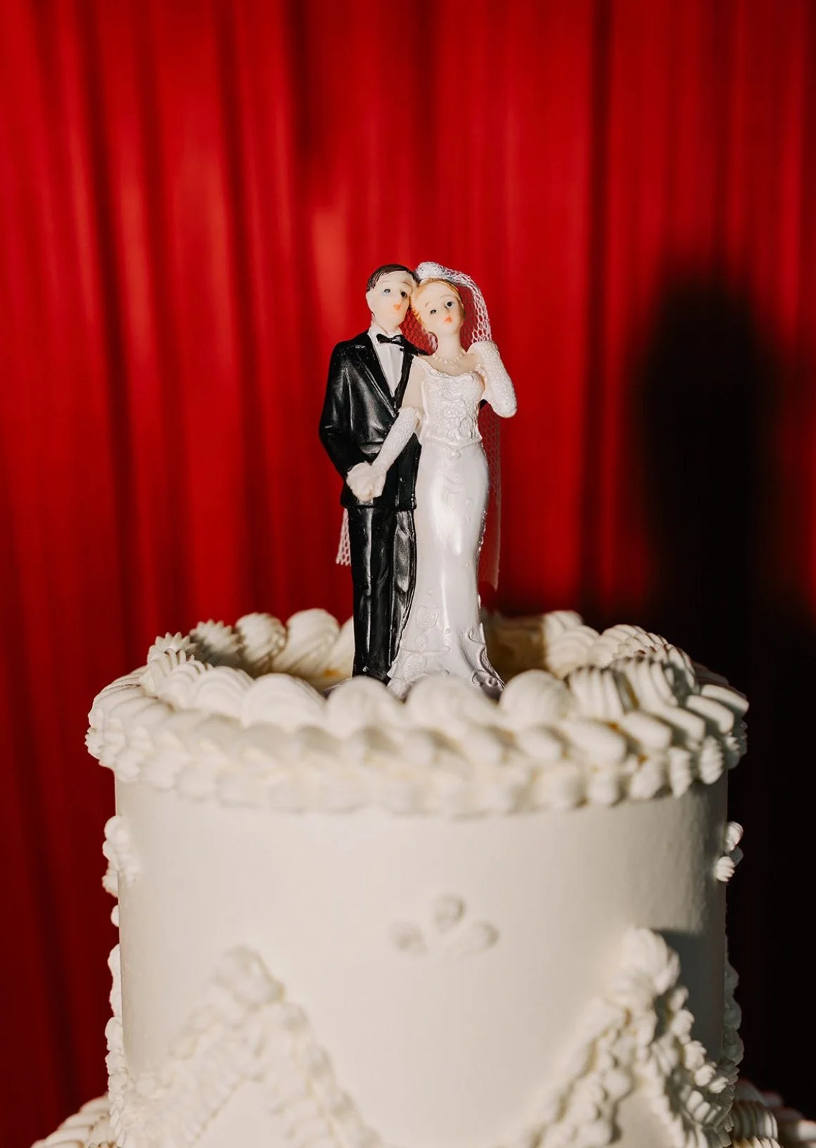 A wedding cake with a bride and groom topper, set against a red curtain background.