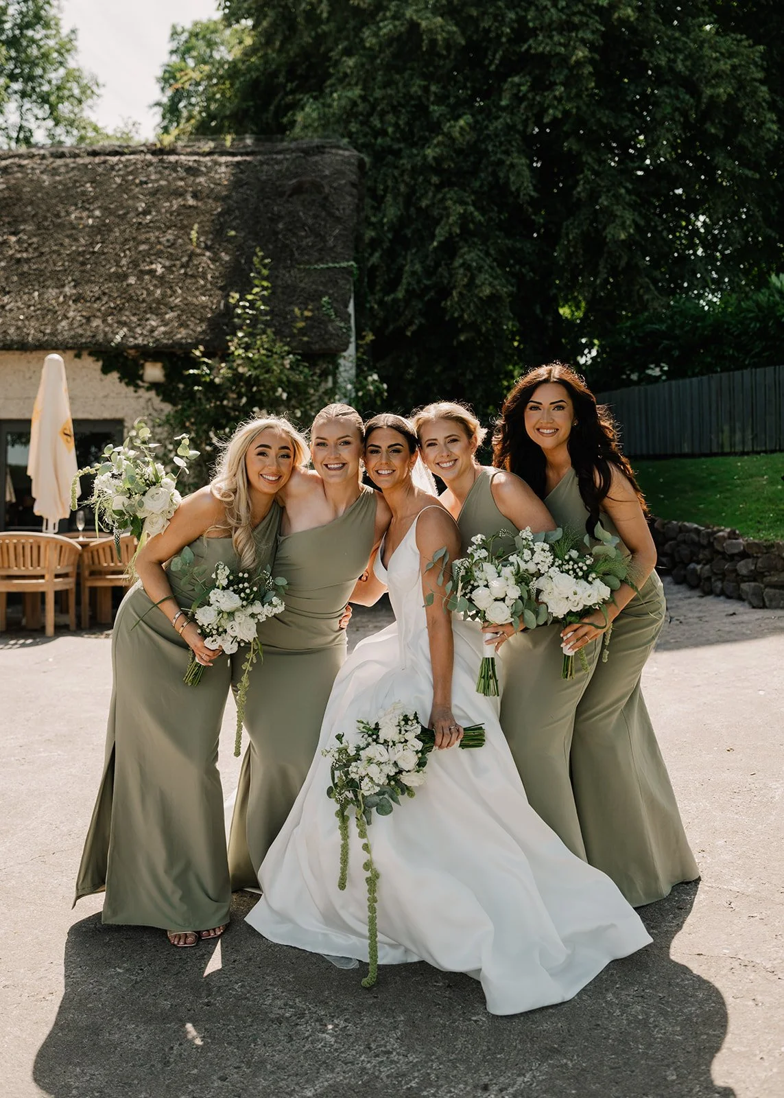 A bride in a white wedding dress surrounded by her four bridesmaids in matching sage green dresses, all holding bouquets of white flowers, posing outdoors in a sunny setting with trees and outdoor furniture in the background.