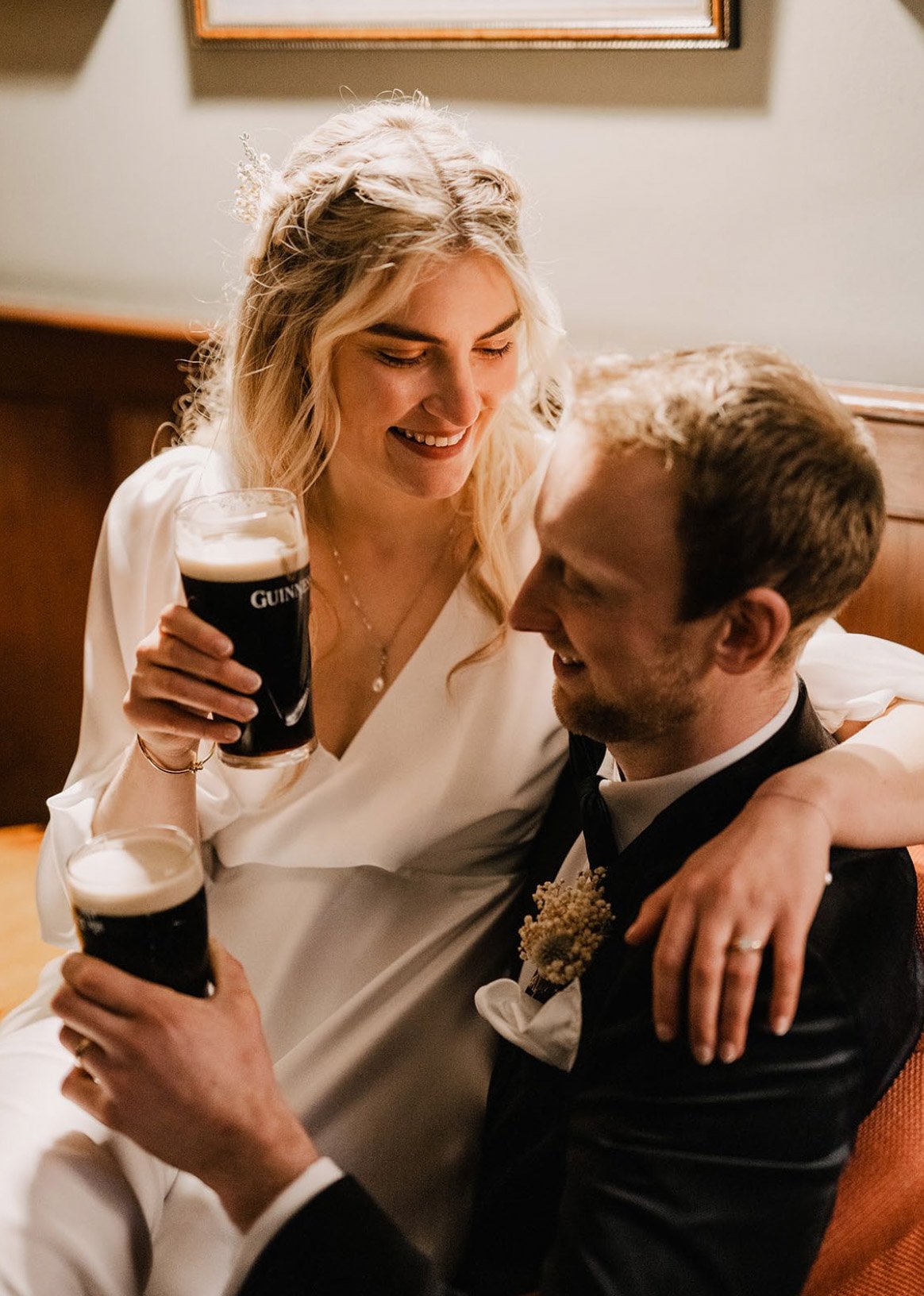 A couple dressed in wedding attire celebrates with pints of Guinness beer. The bride is holding a beer, wearing a white dress, and smiling. The groom is seated, wearing a black suit with a white shirt and a boutonniere, and is looking at the bride with a smile.