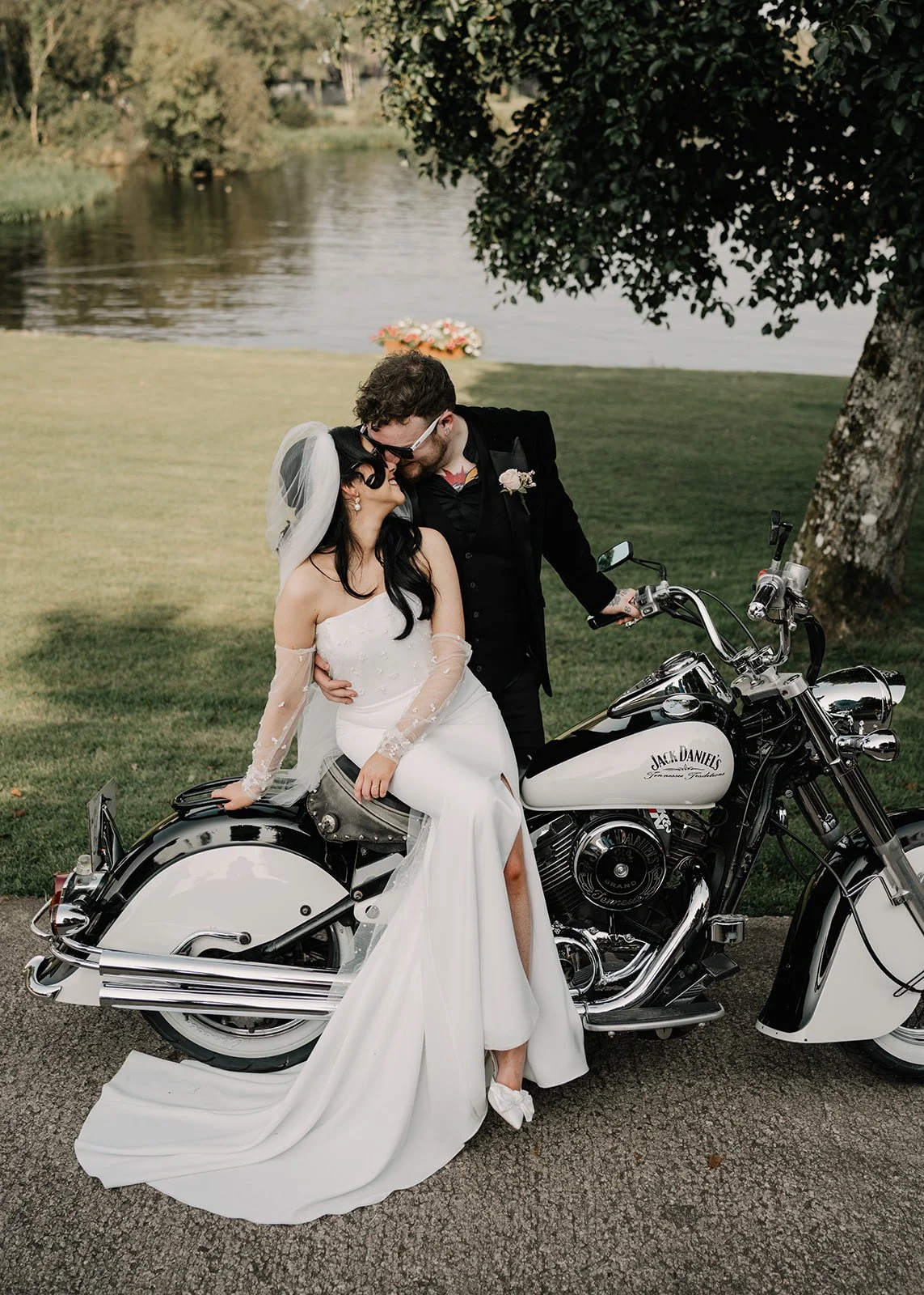 Bride and groom sharing a kiss while sitting on a white motorcycle near a lake, with trees and grass in the background.
