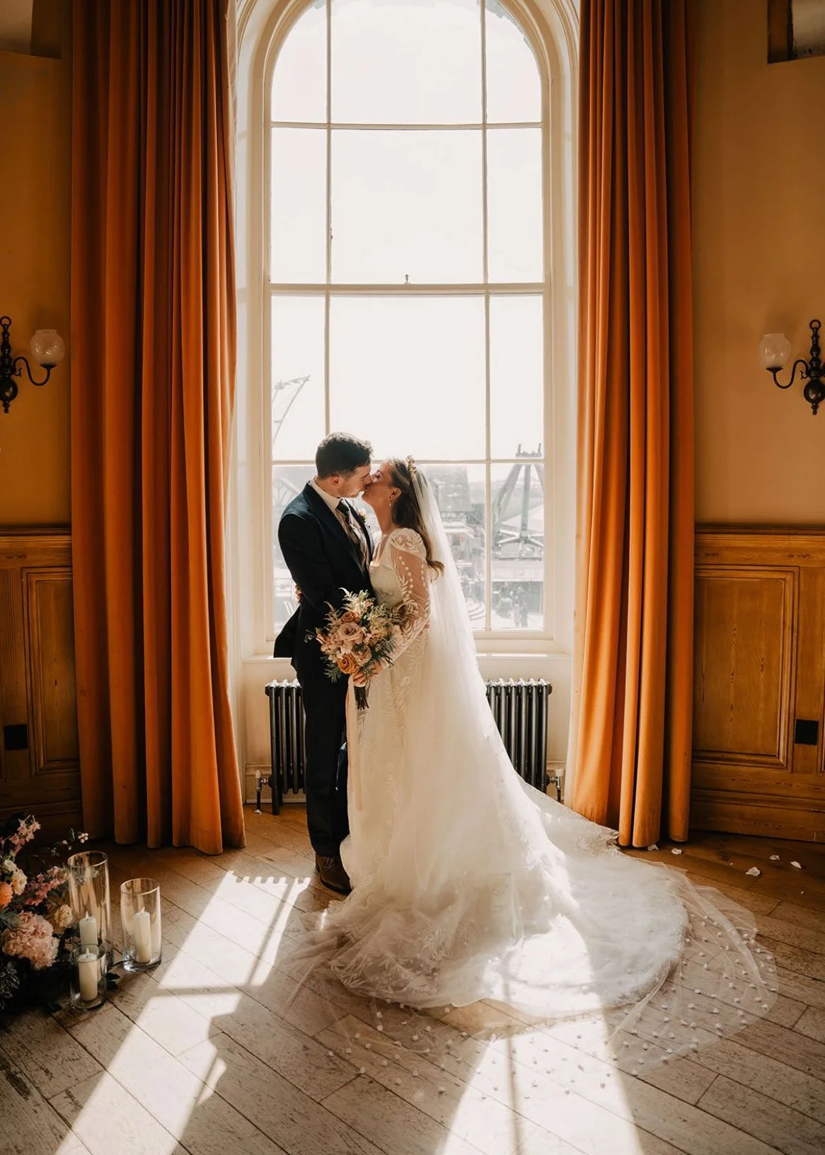 A bride and groom sharing a kiss indoors in front of a large window with orange curtains, with candles and flowers on the floor nearby.
