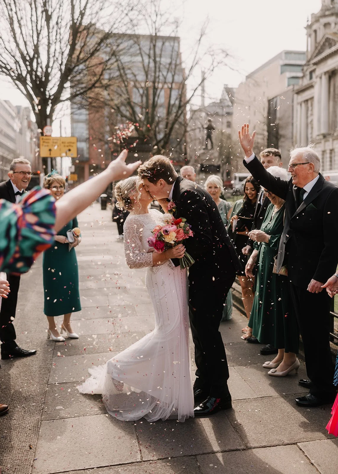 A bride and groom kiss while guests celebrate and throw flower petals on a city sidewalk.