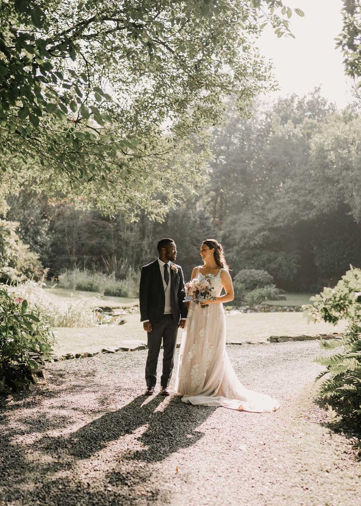 A bride and groom standing together outdoors in a garden, holding hands and smiling at each other, with trees and greenery around them and soft sunlight shining down.