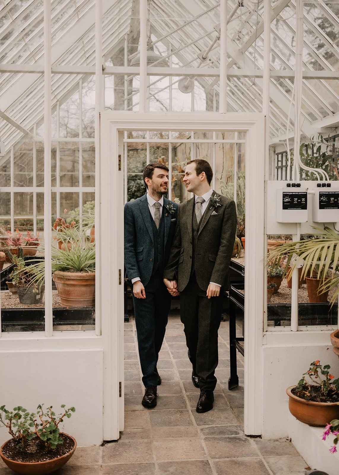 Two men dressed in suits holding hands and smiling at each other inside a greenhouse or conservatory filled with plants.