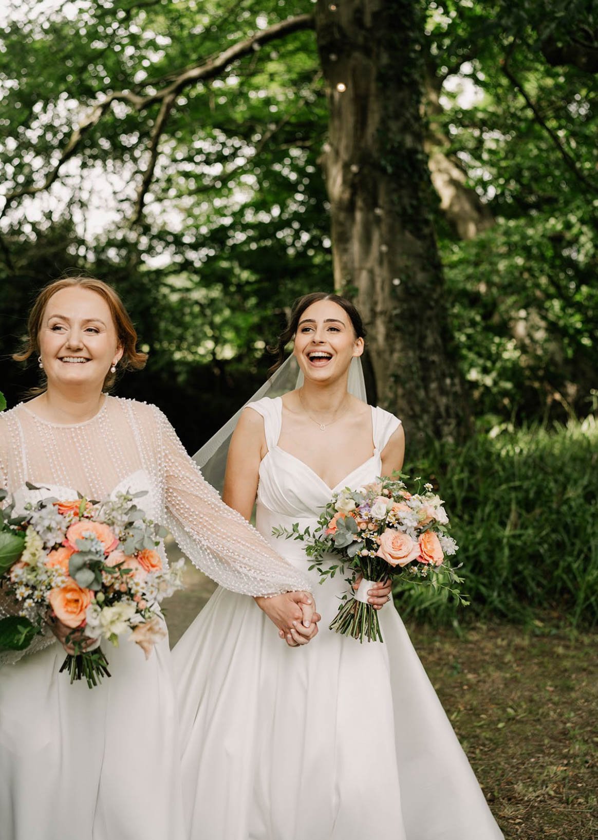 Two women in wedding dresses holding hands and bouquets, smiling and walking outdoors with green foliage and trees in the background.