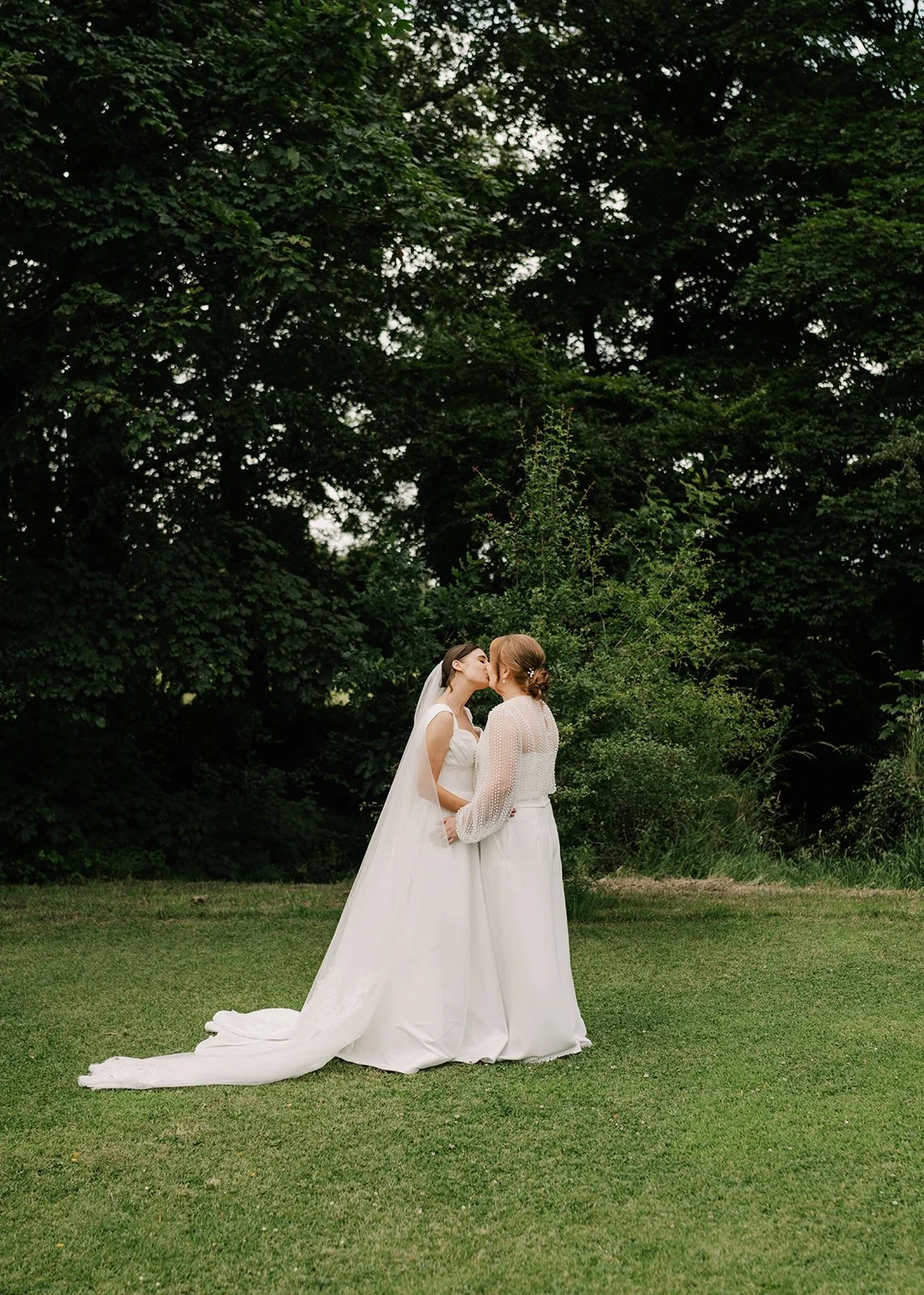 Two women in wedding attire share a kiss outdoors with large trees and greenery in the background.