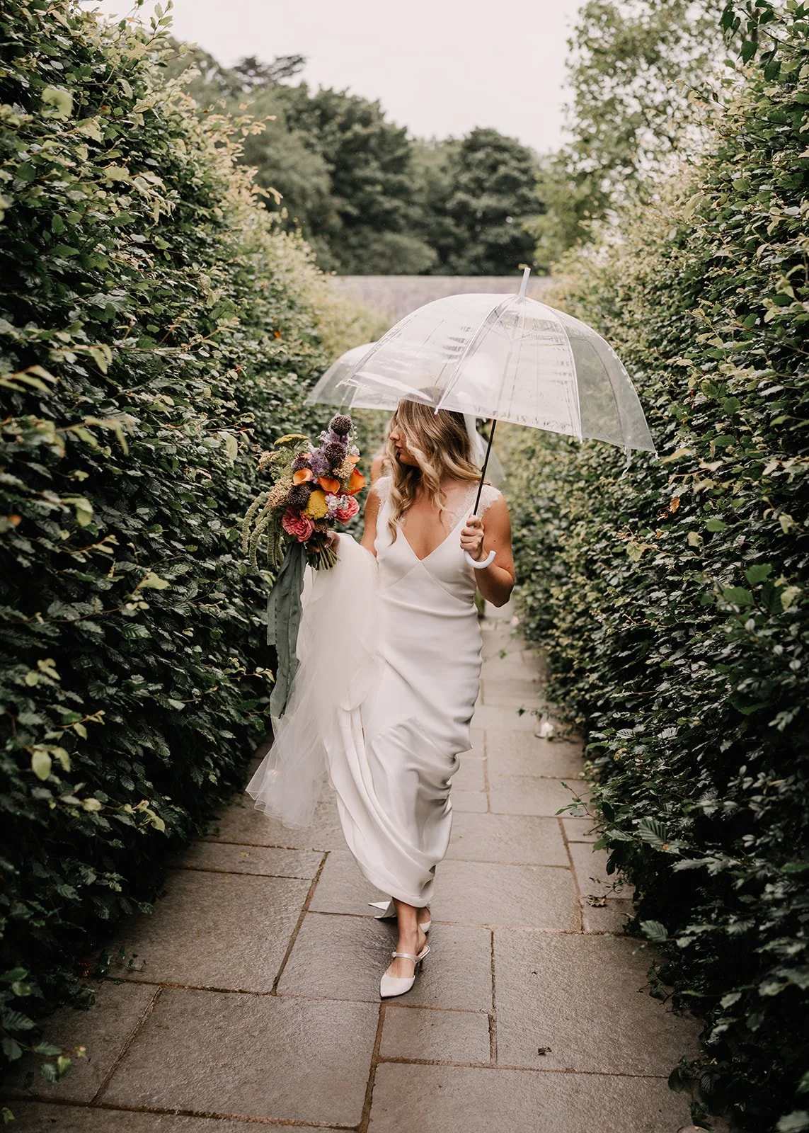 A woman in a white wedding dress holding a bouquet of flowers and an umbrella, walking through a hedge-lined pathway on a rainy day.
