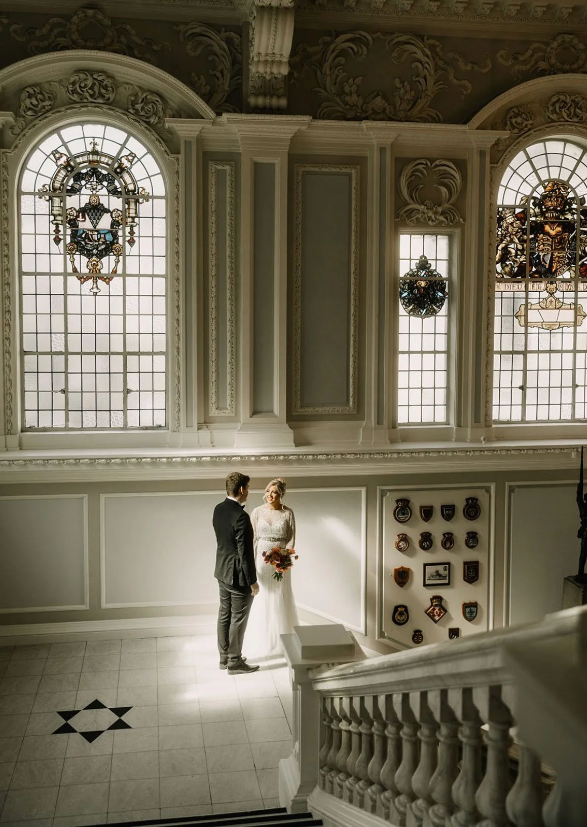A bride in a white wedding dress holding a bouquet and a groom in a black suit standing in a grand, ornate hall with large stained glass windows and decorative moldings, engaged in a moment of conversation.