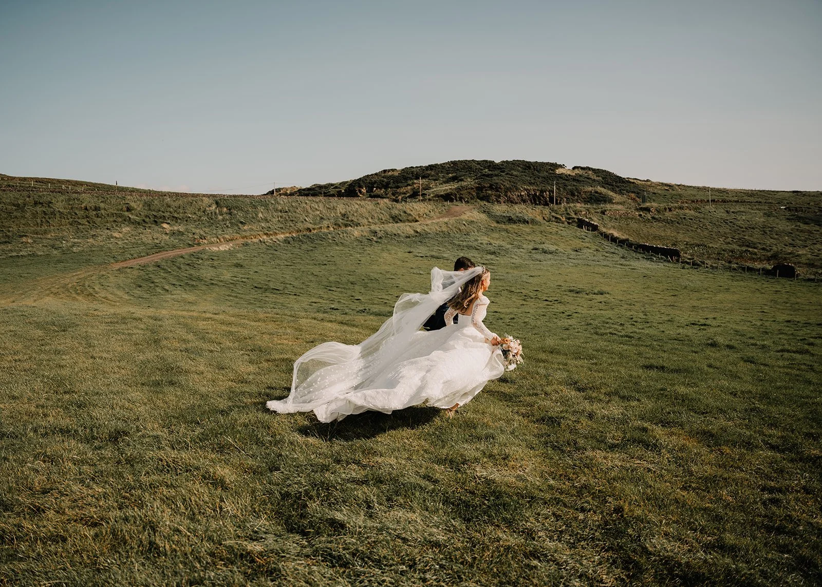 Bride in a white wedding dress holding a bouquet running across a grassy field.