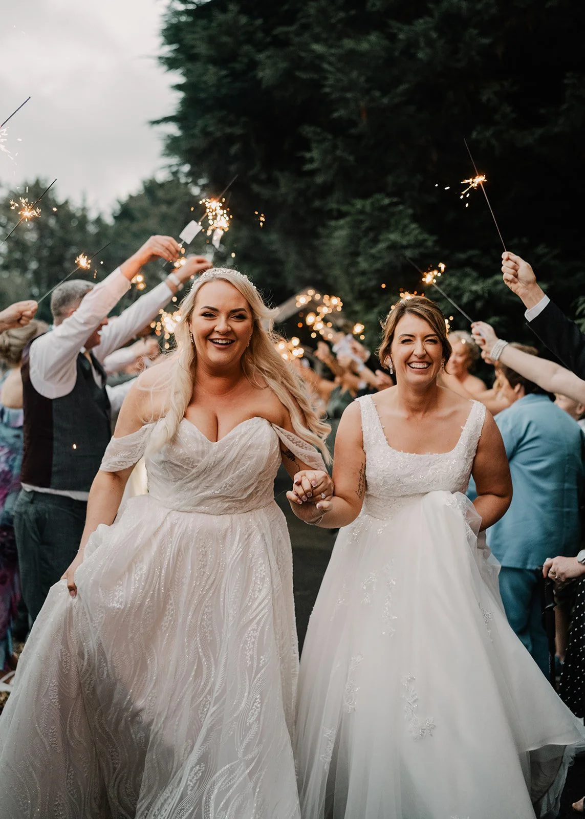 Two women in wedding dresses holding hands and smiling, surrounded by friends and family holding sparklers outside with trees in the background.