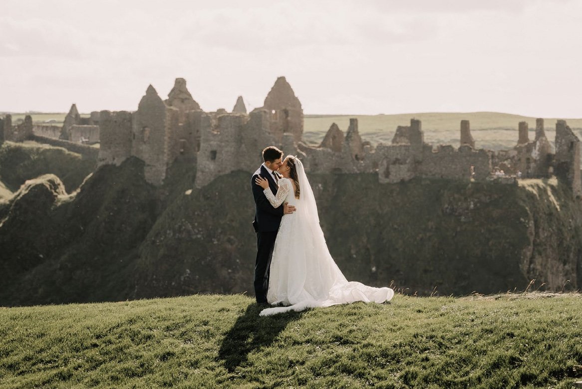 A bride and groom embrace outdoors with a castle in the background.