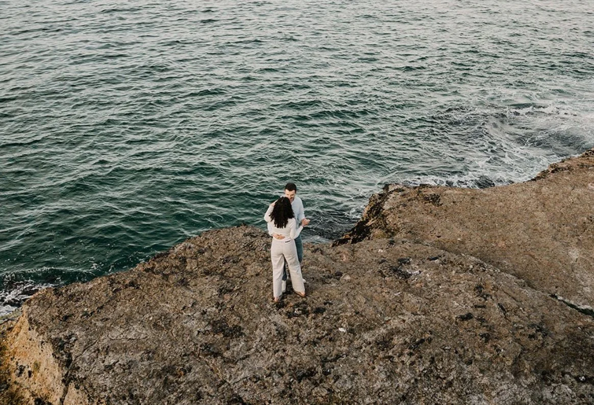 A couple standing on a rocky shore near the ocean, facing each other and holding hands.