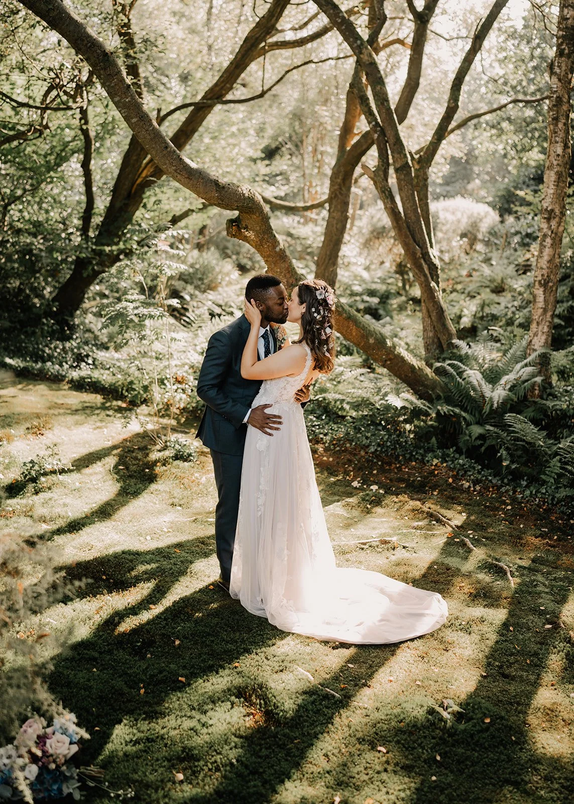 A bride and groom sharing a kiss in a lush, sunlit forest during their wedding.