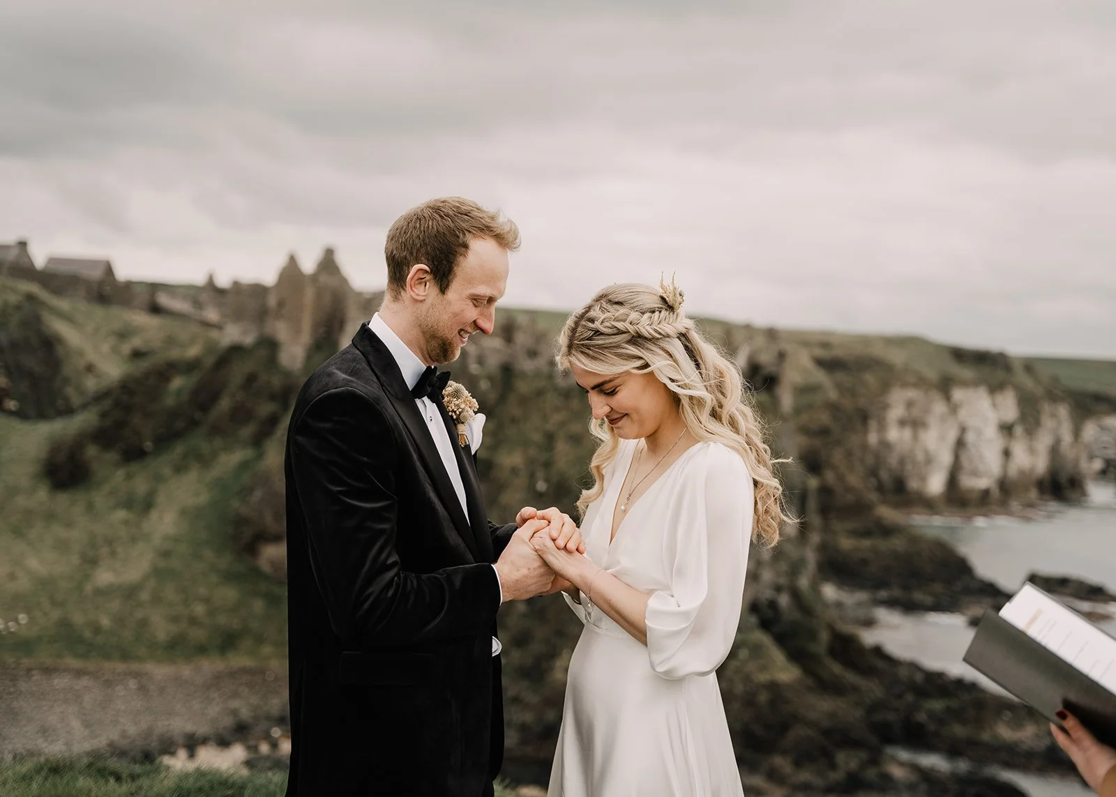 A couple getting married outdoors near the coastline, with cliffs and ruins in the background. The groom is wearing a tuxedo and the bride a white dress, both smiling and holding hands.