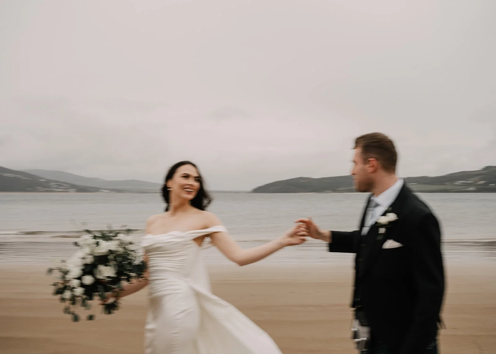 A bride and groom holding hands and dancing on a beach near a body of water with mountains in the background.
