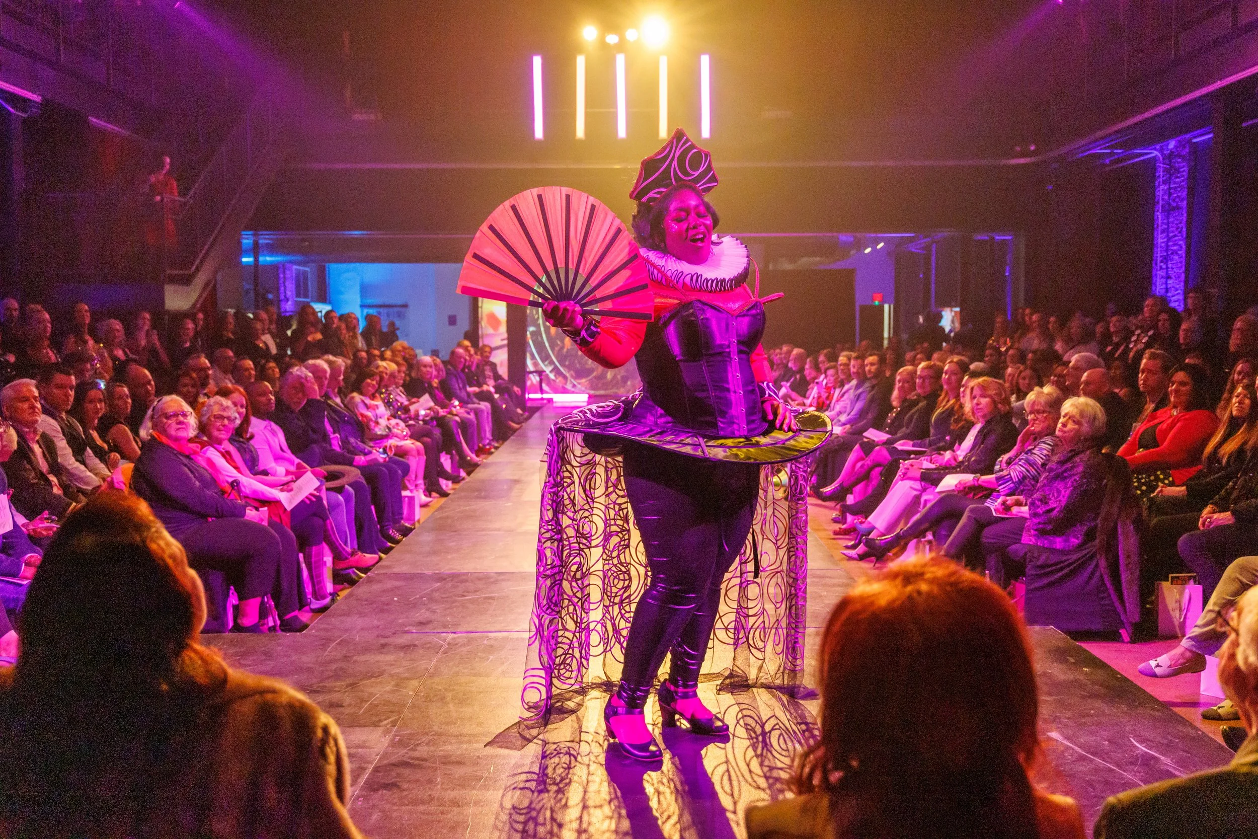 Performer on stage wearing colorful, avant-garde costume holding a fan, audience watching in a dark venue with vibrant stage lighting.