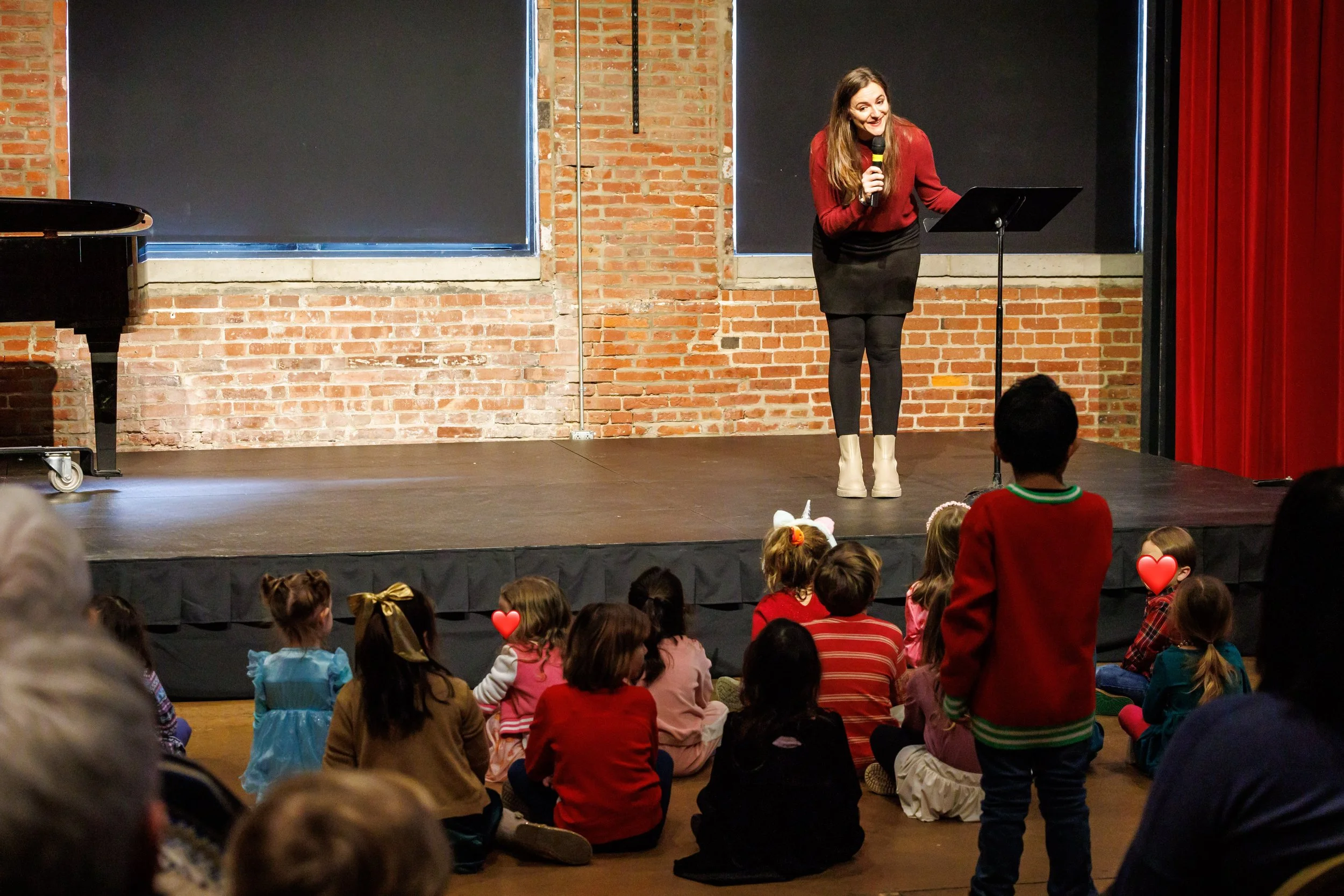 A woman on stage holding a microphone, performing in front of an audience of children sitting on the floor. The stage has a brick wall background and a red curtain on the side.
